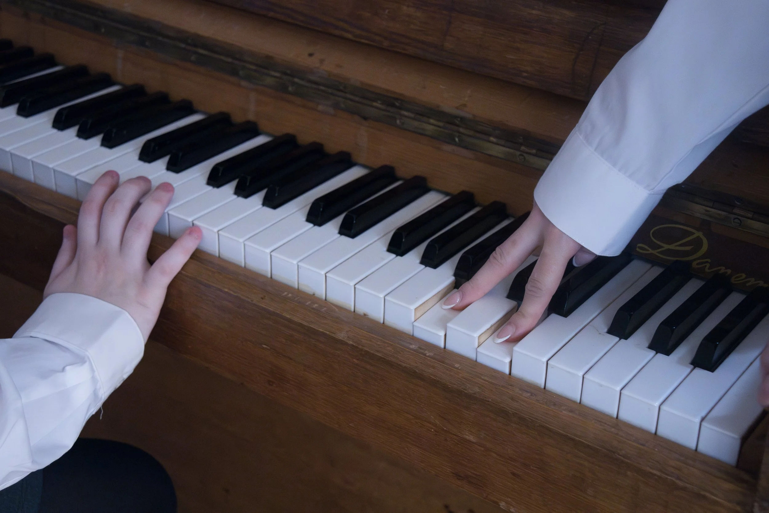 A close-up of a person's hands playing a wooden piano with black and white keys. The person is wearing a white long-sleeve shirt, and the background shows a wooden surface.