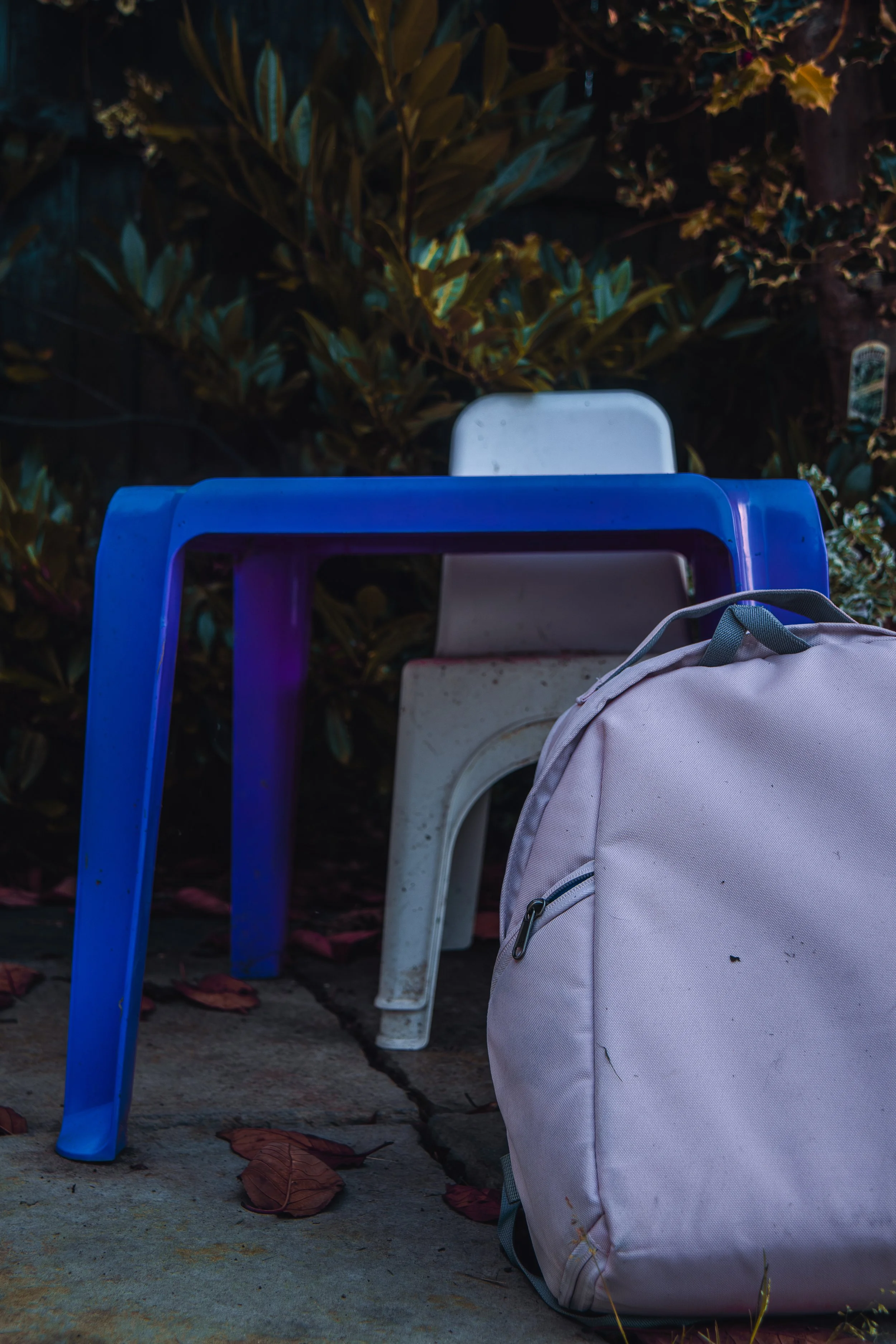 A white backpack resting on the ground next to a purple outdoor table and a white plastic chair in a garden at night, with leaves and a fence in the background.
