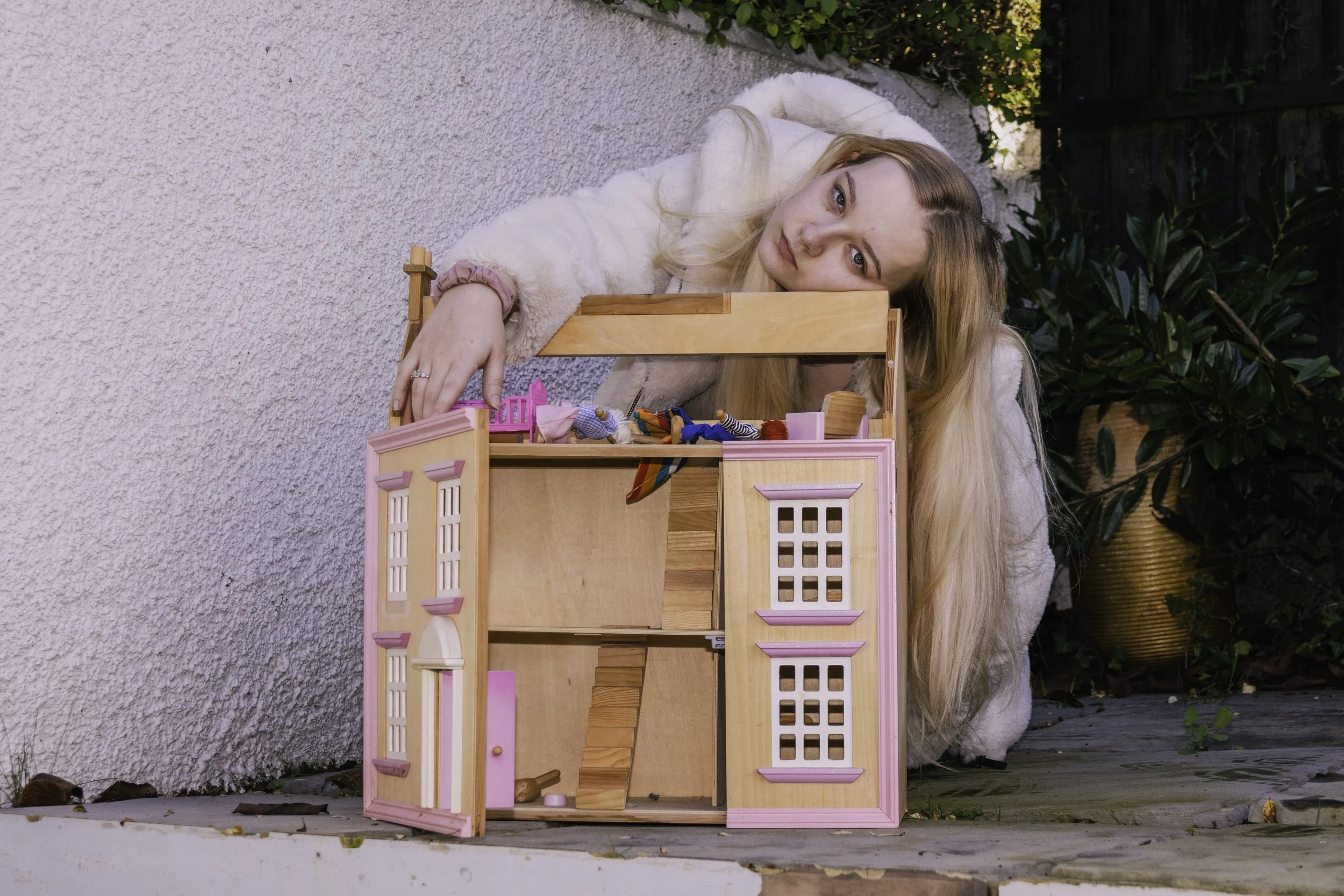 A young woman with long blonde hair lying on her side outdoors next to a pink dollhouse on a wooden deck, surrounded by greenery and plants.
