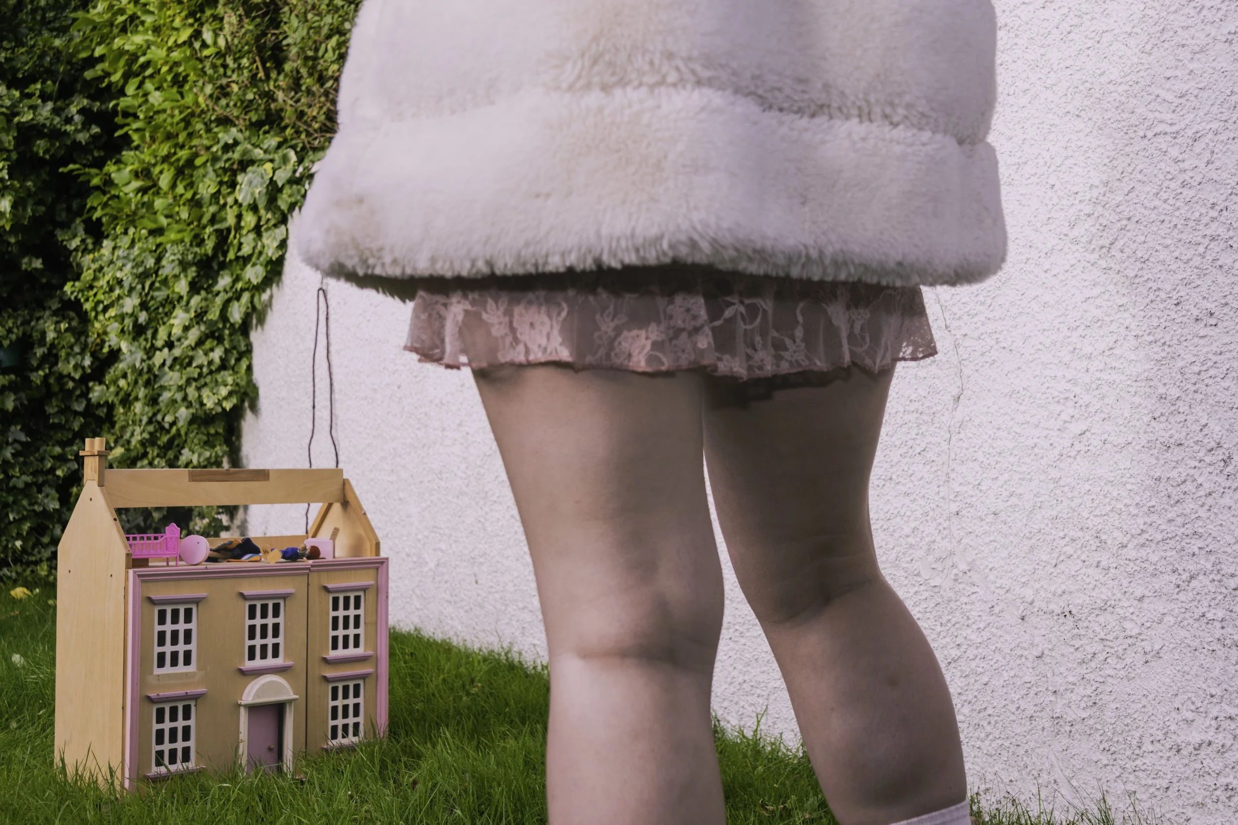 Close-up of a woman's lower back and legs, wearing pink lace shorts and a fluffy pink jacket, standing on grass outside near a white wall with a small pink and beige dollhouse and toys nearby.