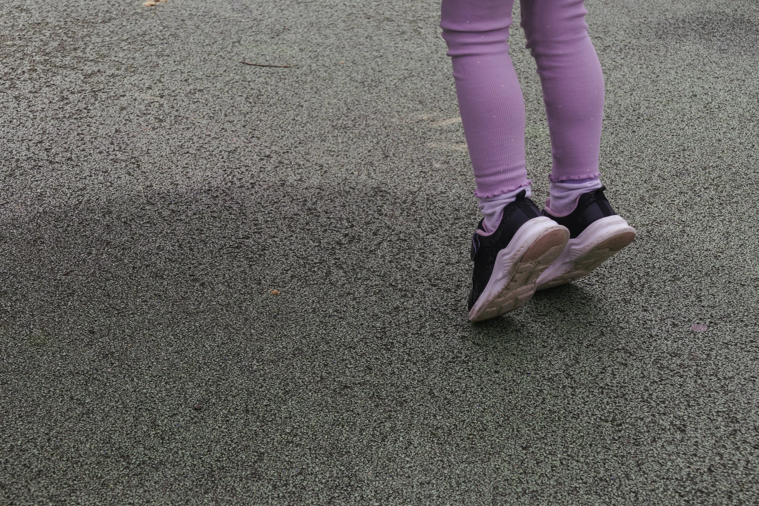Close-up of a child's legs and feet standing on a textured asphalt surface, wearing purple leggings and black sneakers with white soles.