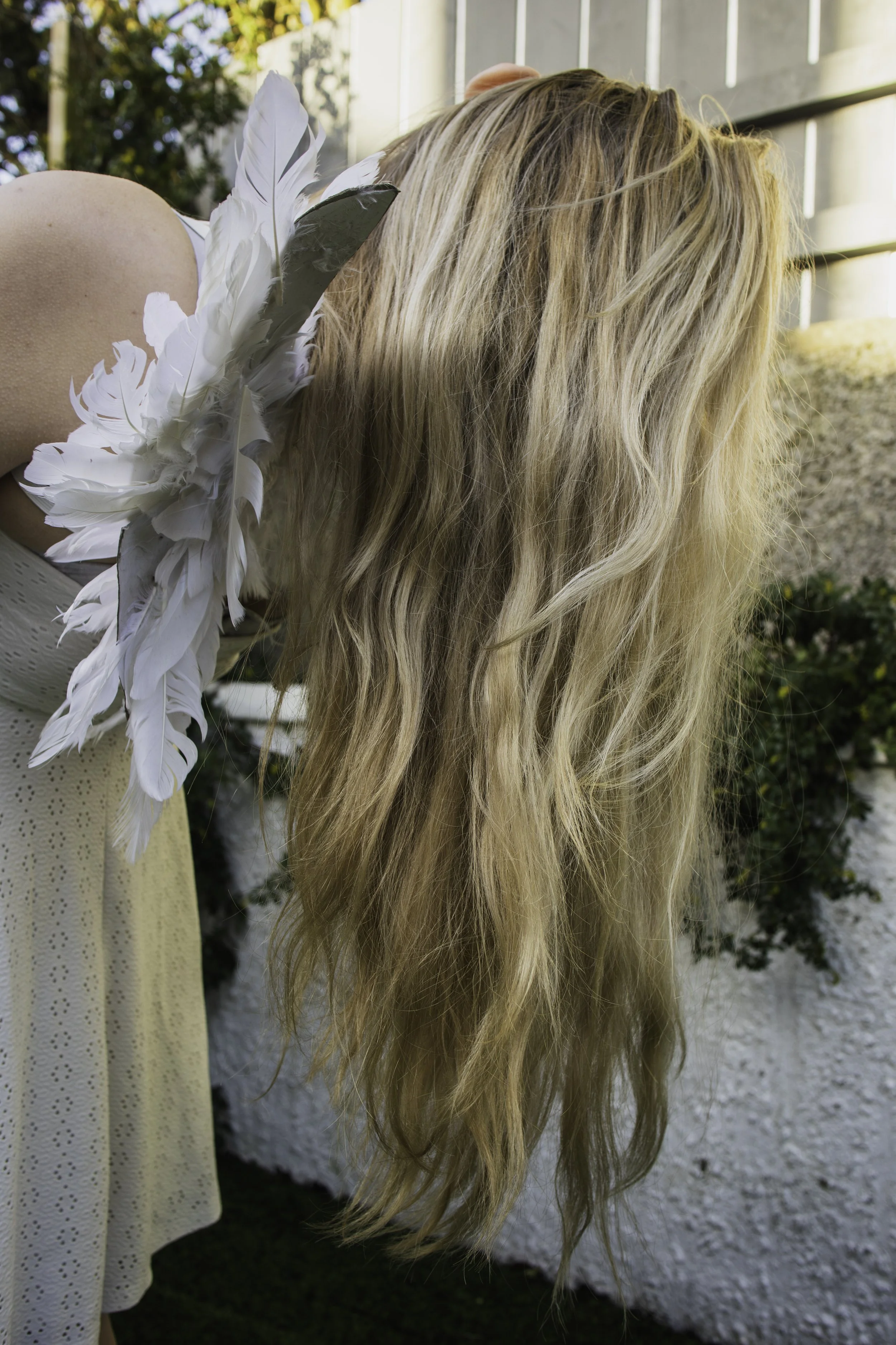 Close-up of a woman with long, wavy blonde hair wearing white feathered earrings and a white flowy dress, outdoors near a white wall and greenery.
