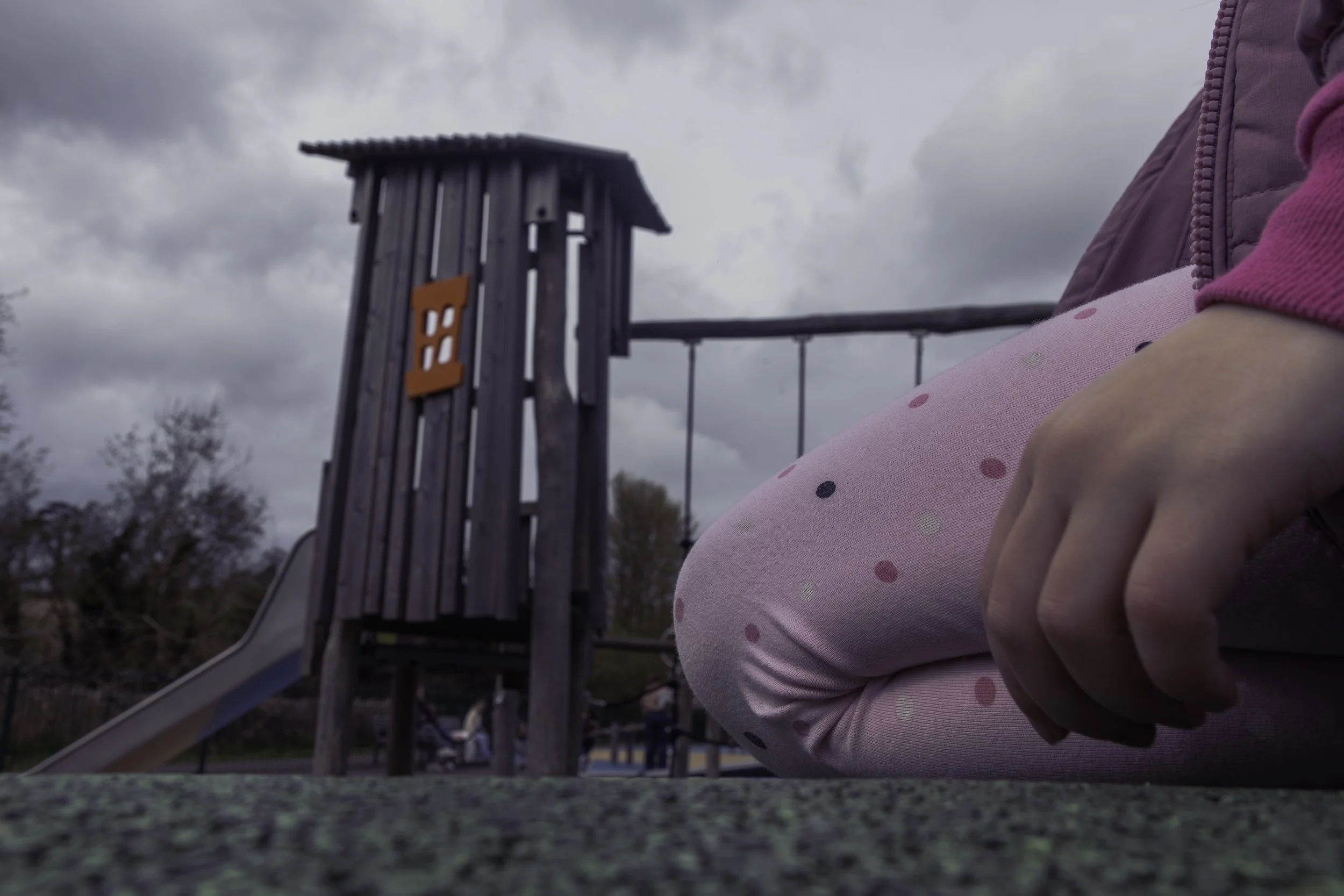 Close-up of children holding hands, with a wooden playground structure in the background, against a cloudy sky.
