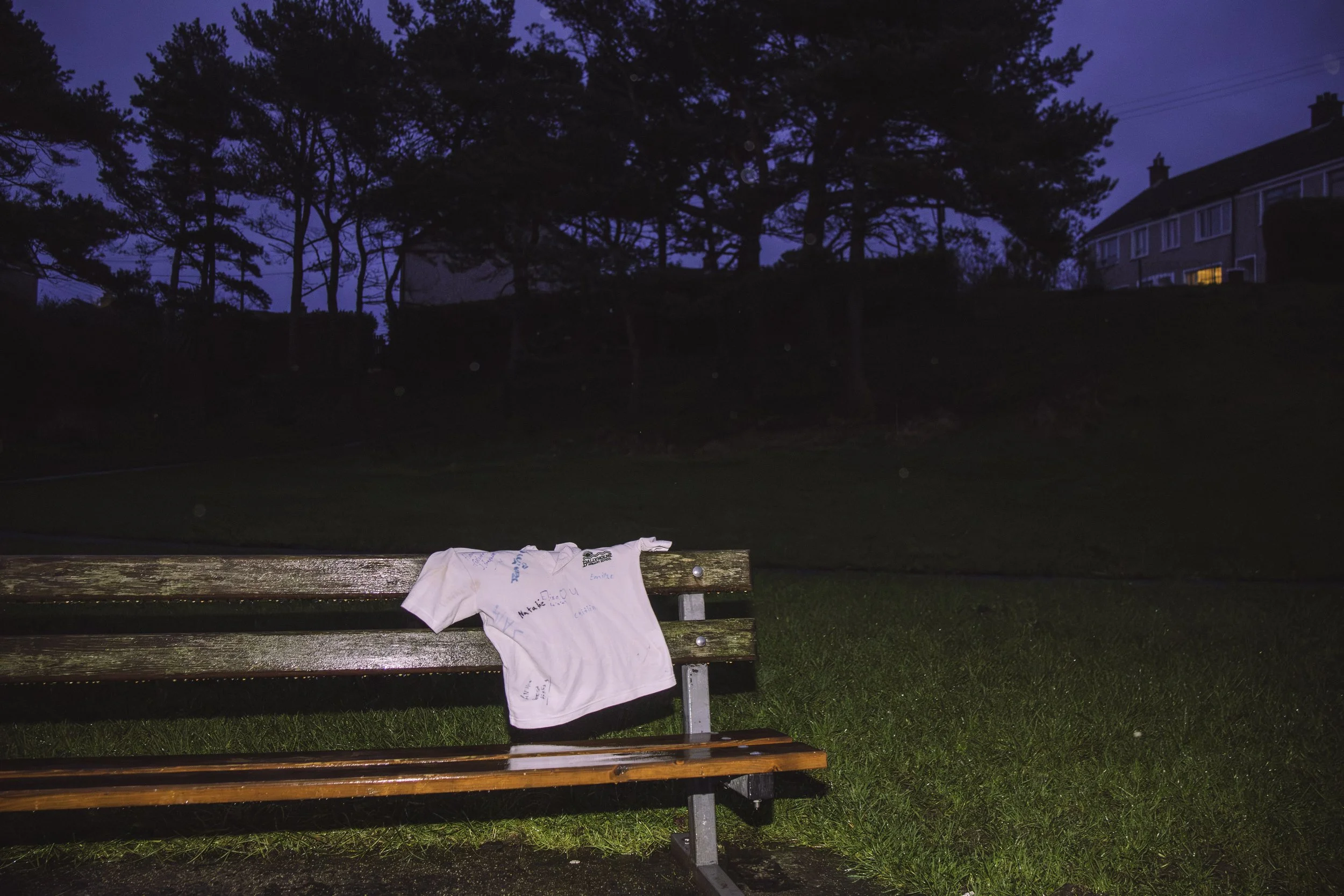 A park bench with a small polo shirt draped over it, set against a dark evening background with trees and houses visible in the distance.