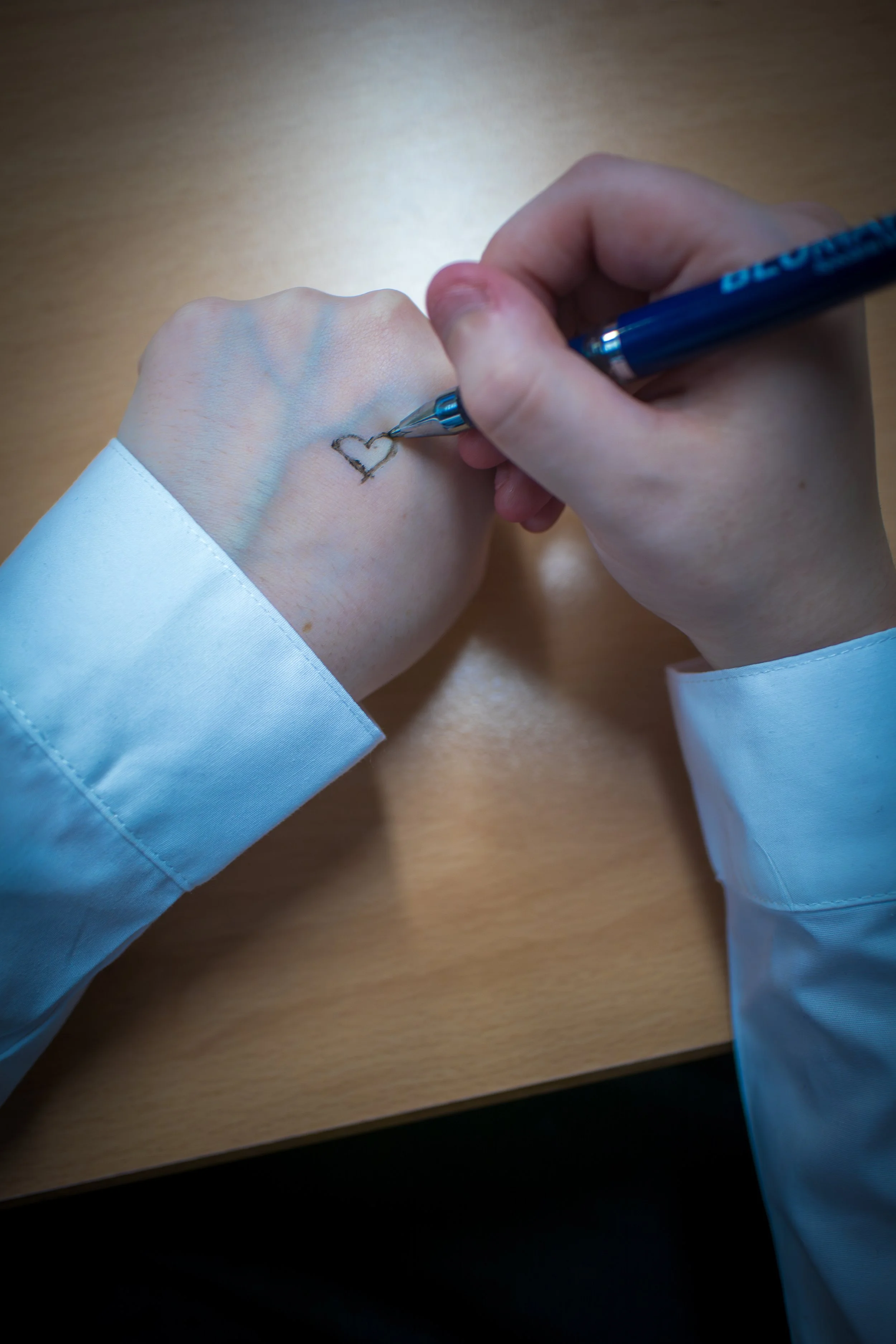 Person drawing a heart on the back of another person's hand with a blue pen, wearing a white long-sleeve shirt.