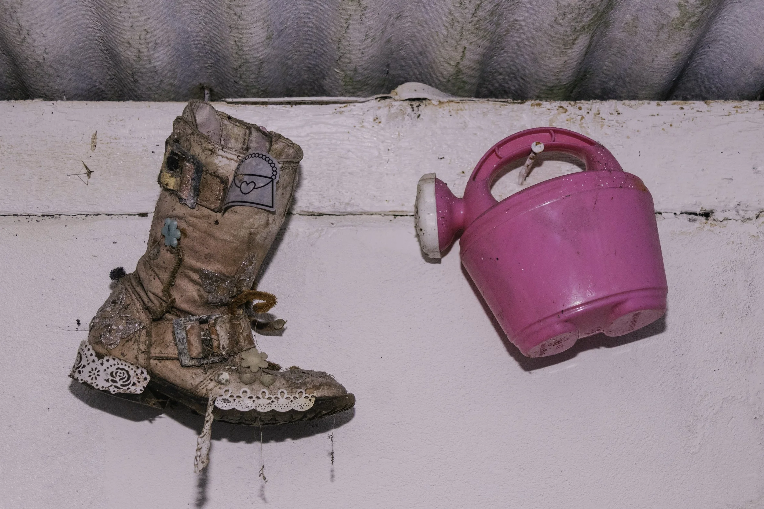 A pink plastic toy watering can and a well-worn children's shoe on a white painted surface.