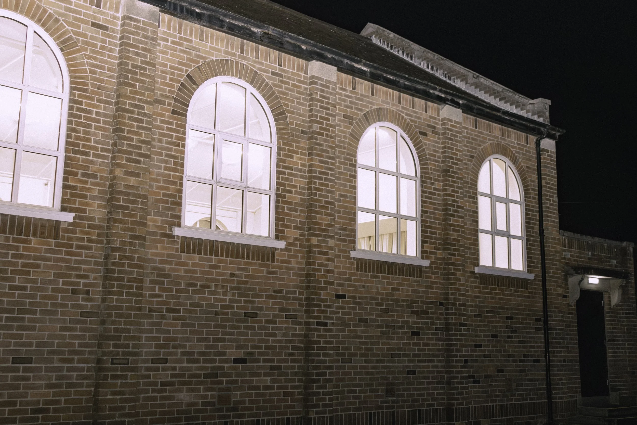 Nighttime view of a brick building with multiple large arched windows illuminated from inside. The building features brick arches above each window and a black gutter running along the roof's edge. Part of the dark night sky is visible above the buil