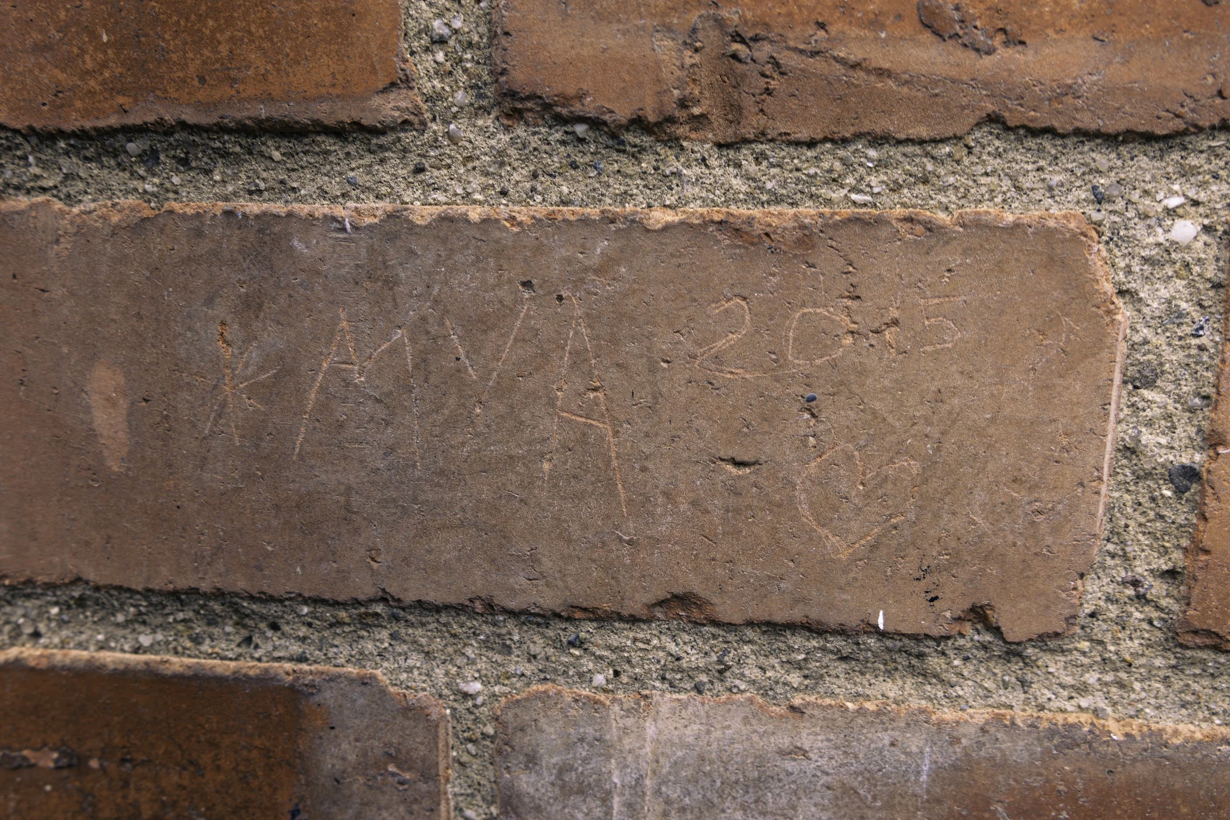 Close-up of red brick wall with graffiti etched into the bricks reading 'Mya 2015'.