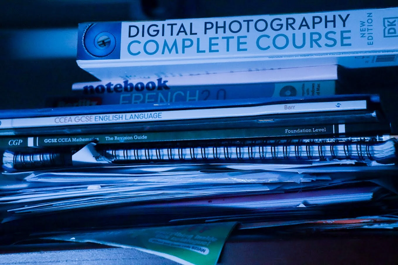A collection of textbooks, notebooks, and papers stacked on a shelf, including a 'Digital Photography Complete Course' book, a notebook, and various study guides for GCSE and foundation level exams.