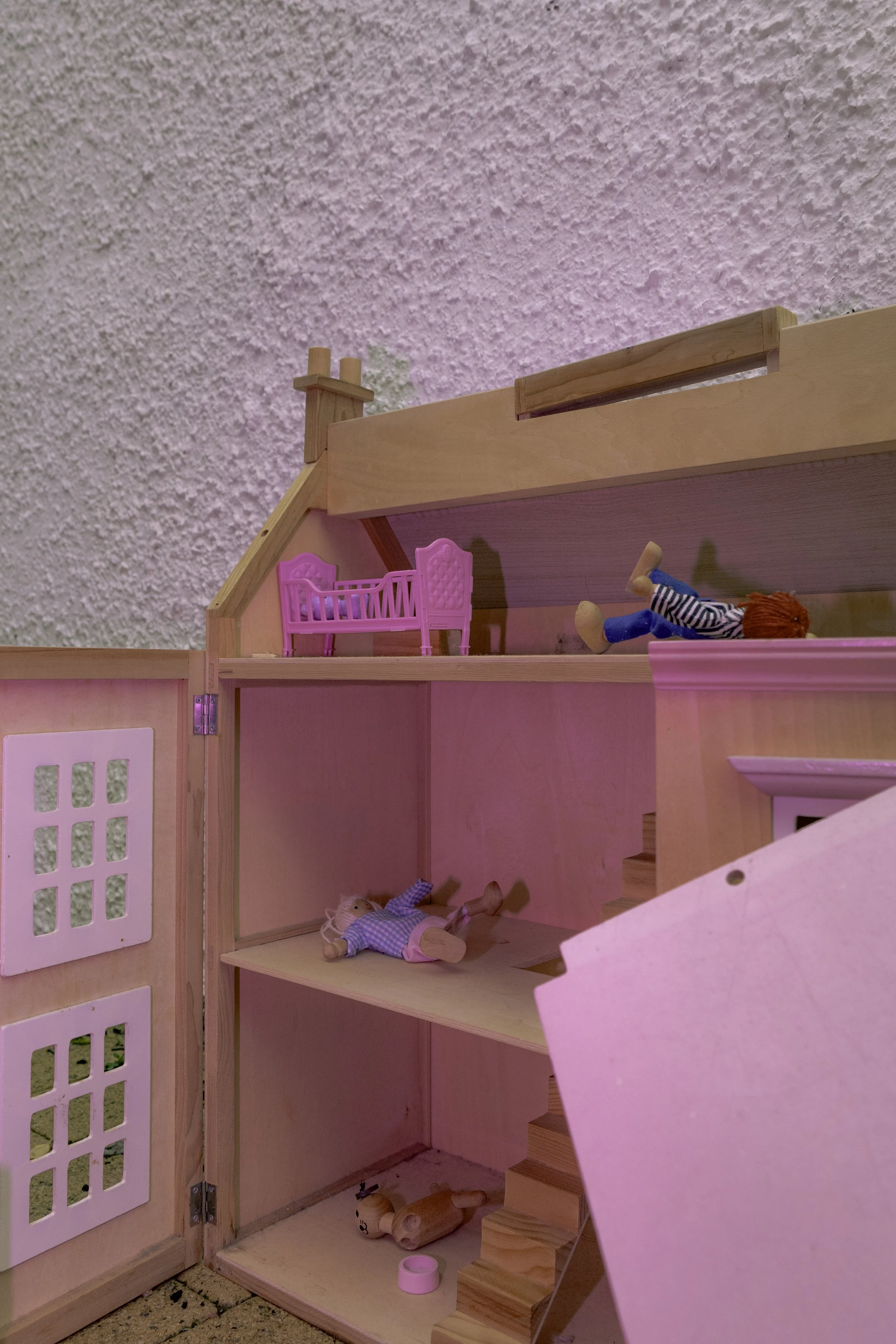 A dollhouse bedroom with pink walls, wooden stairs, and furniture. There is a doll with red hair lying on the bed and another doll with blonde hair lying on the floor.