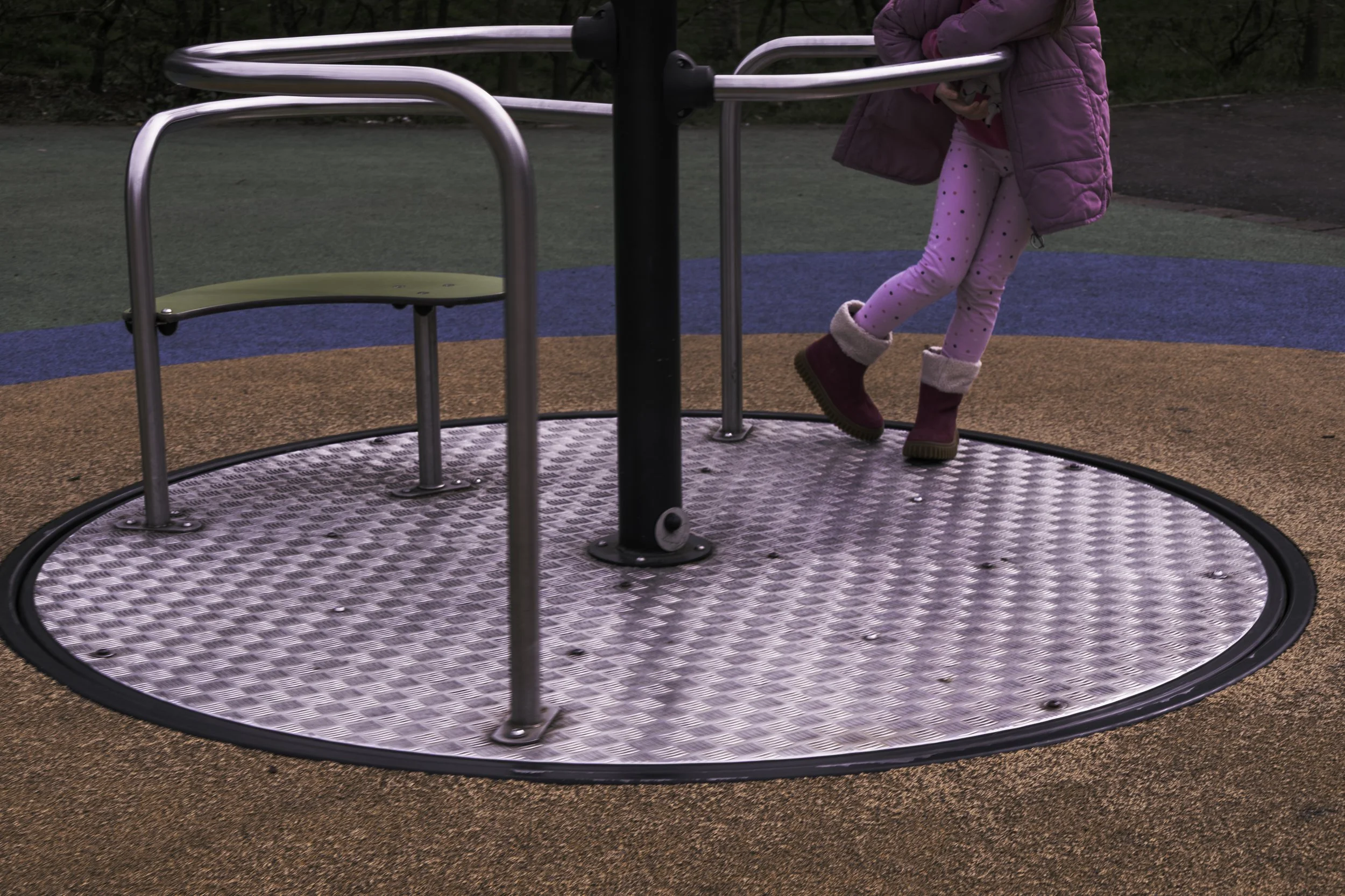 A young girl wearing a pink jacket and pink polka-dot leggings standing on a playground merry-go-round with metal bars and a textured metal surface, surrounded by a colorful asphalt surface and trees in the background.