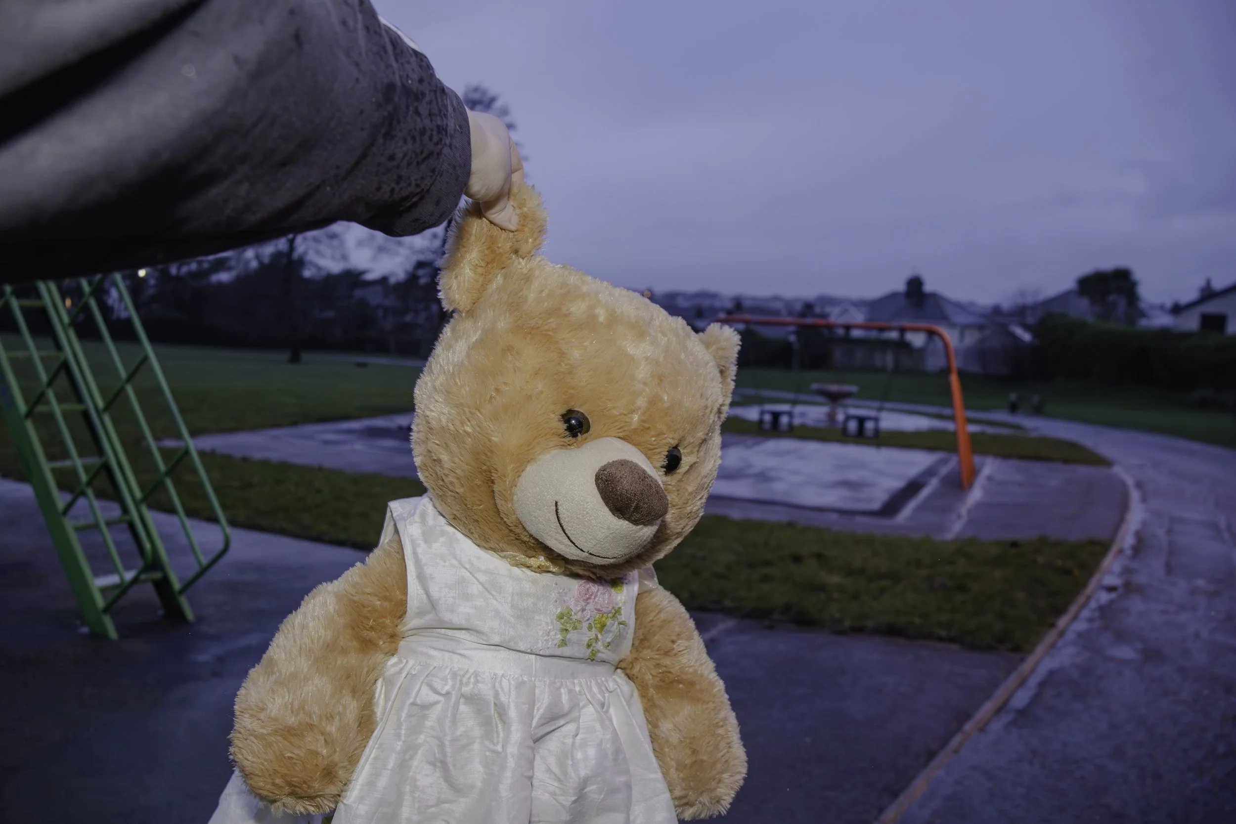 A teddy bear dressed in a white dress standing outdoors in a park during dusk, with playground equipment in the background.
