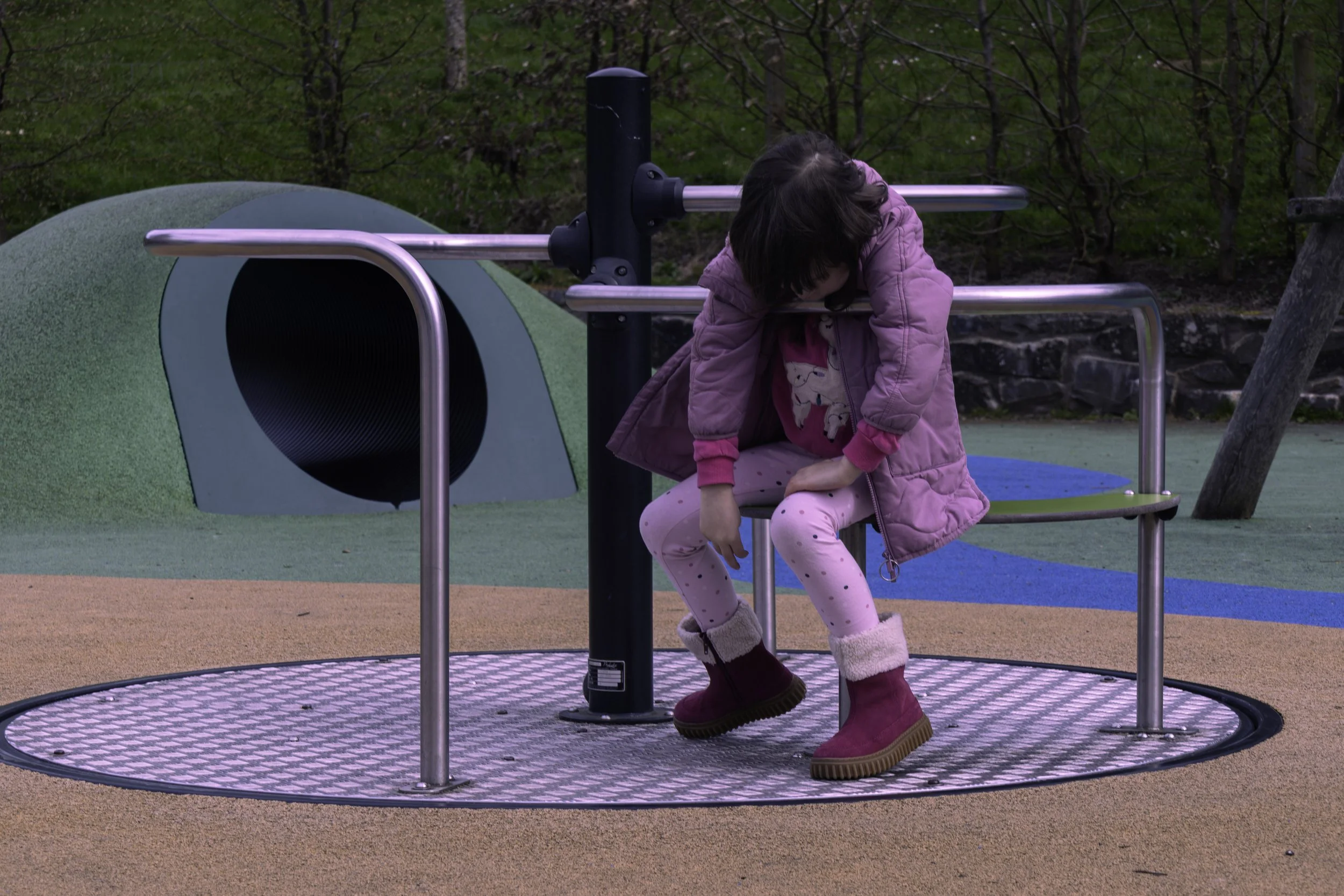 A young girl wearing a pink coat, pink pajamas with unicorns, and pink boots sitting on a round play apparatus outdoors at a playground, with green trees and colorful ground surface in the background.