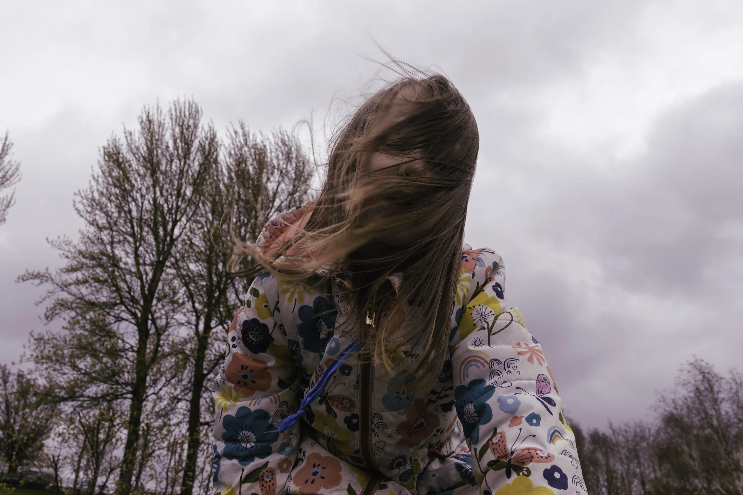 A girl with long hair blowing in the wind, wearing a colorful floral jacket, standing outdoors with trees and a cloudy sky in the background.