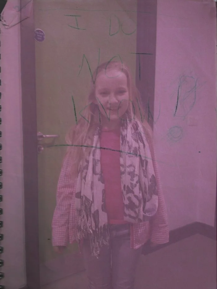 A young girl smiling with writing on it that says, "I do not know!" The image has a pink tint, possibly due to lighting or a filter. The girl is wearing a checkered jacket and a scarf, standing in a hallway next to a clincal door.