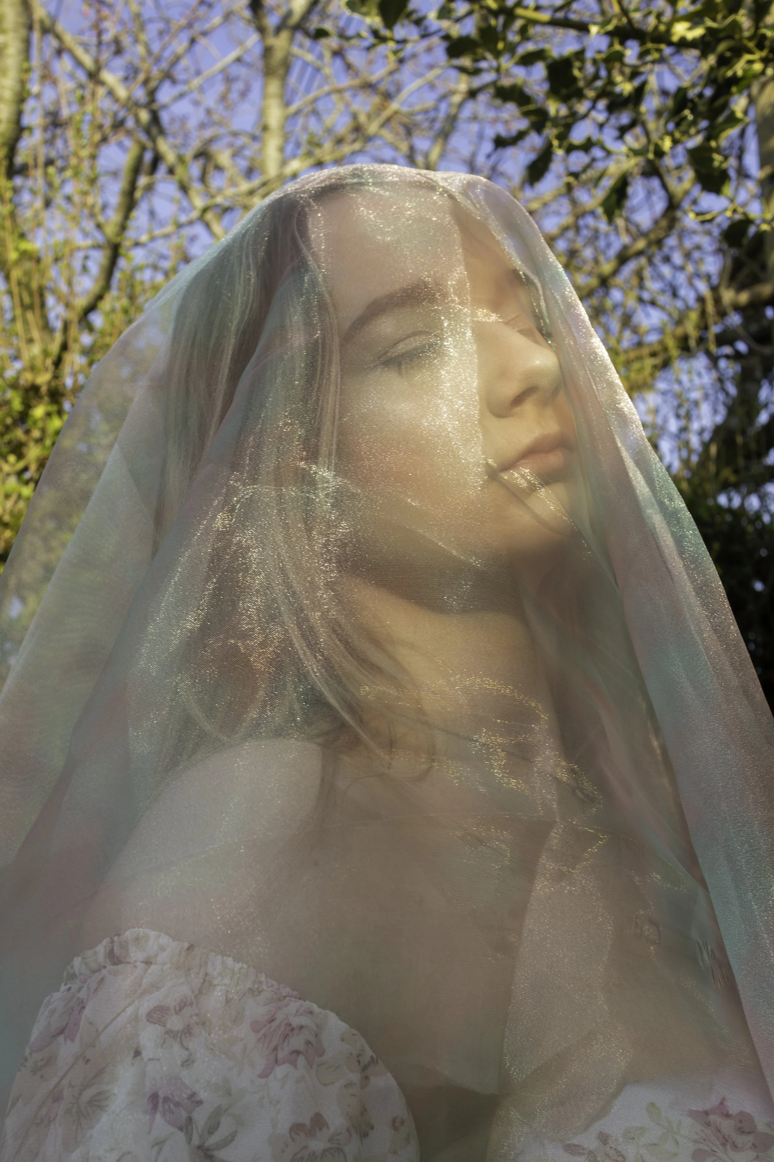 A woman with closed eyes and a gentle smile, partially covered with a translucent, shimmering fabric, outdoors with trees and blue sky in the background.
