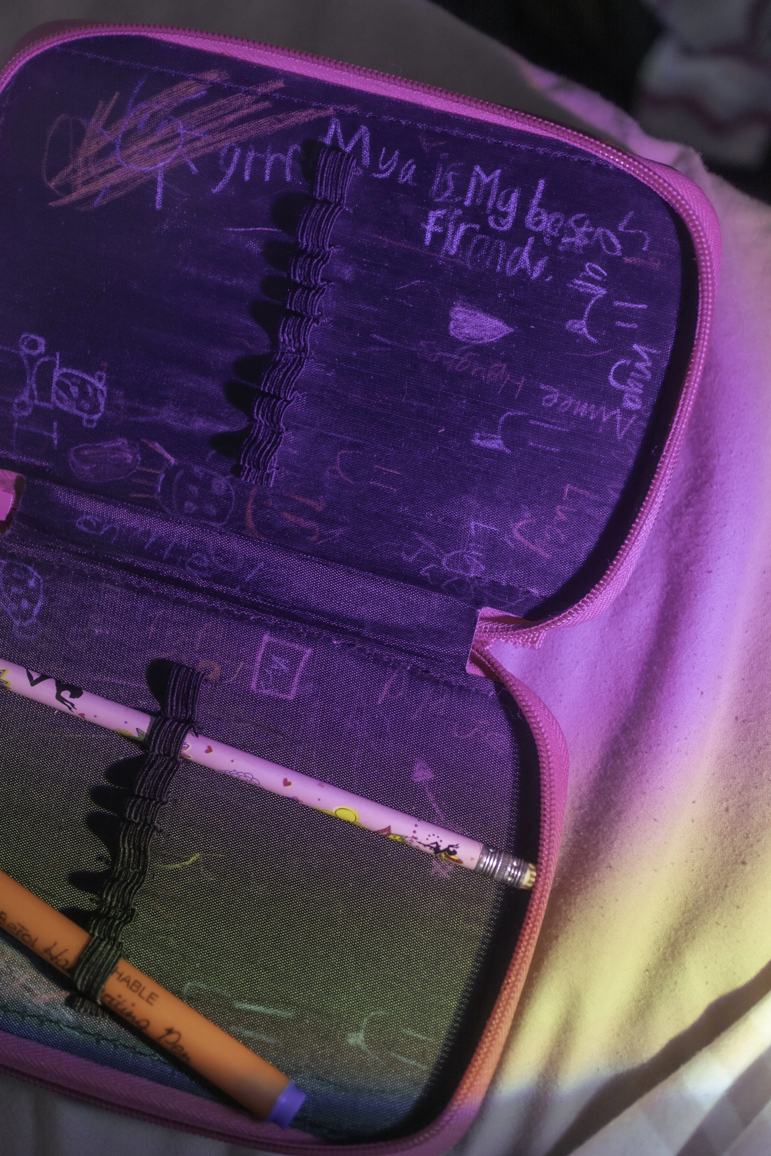 A pencilcase with pink zipper and black mesh pocket containing colorful pens, scribbled notes, and doodles.