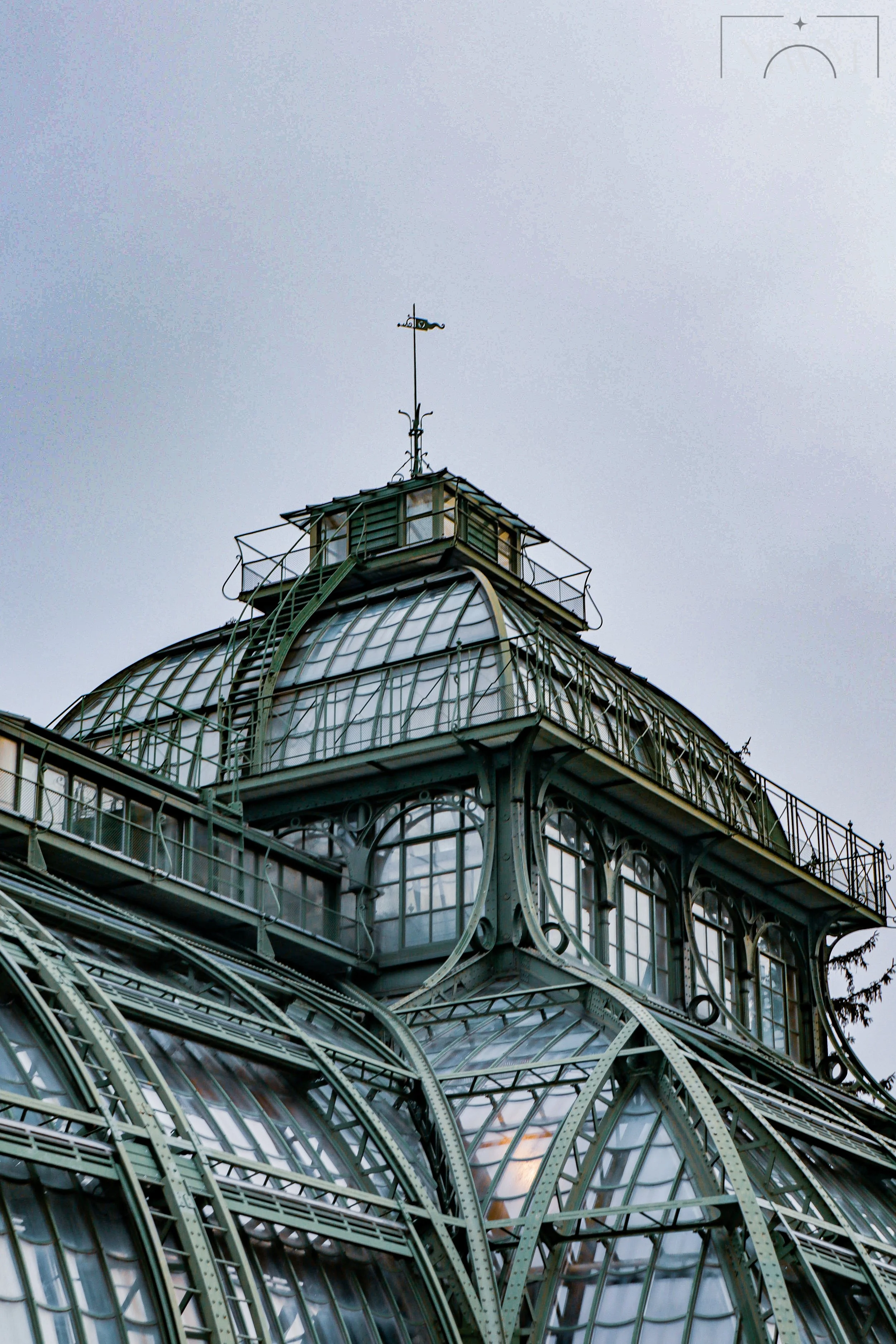Close-up of a historic glass and metal-domed building with intricate architectural details and a weather vane on top, set against a cloudy sky.