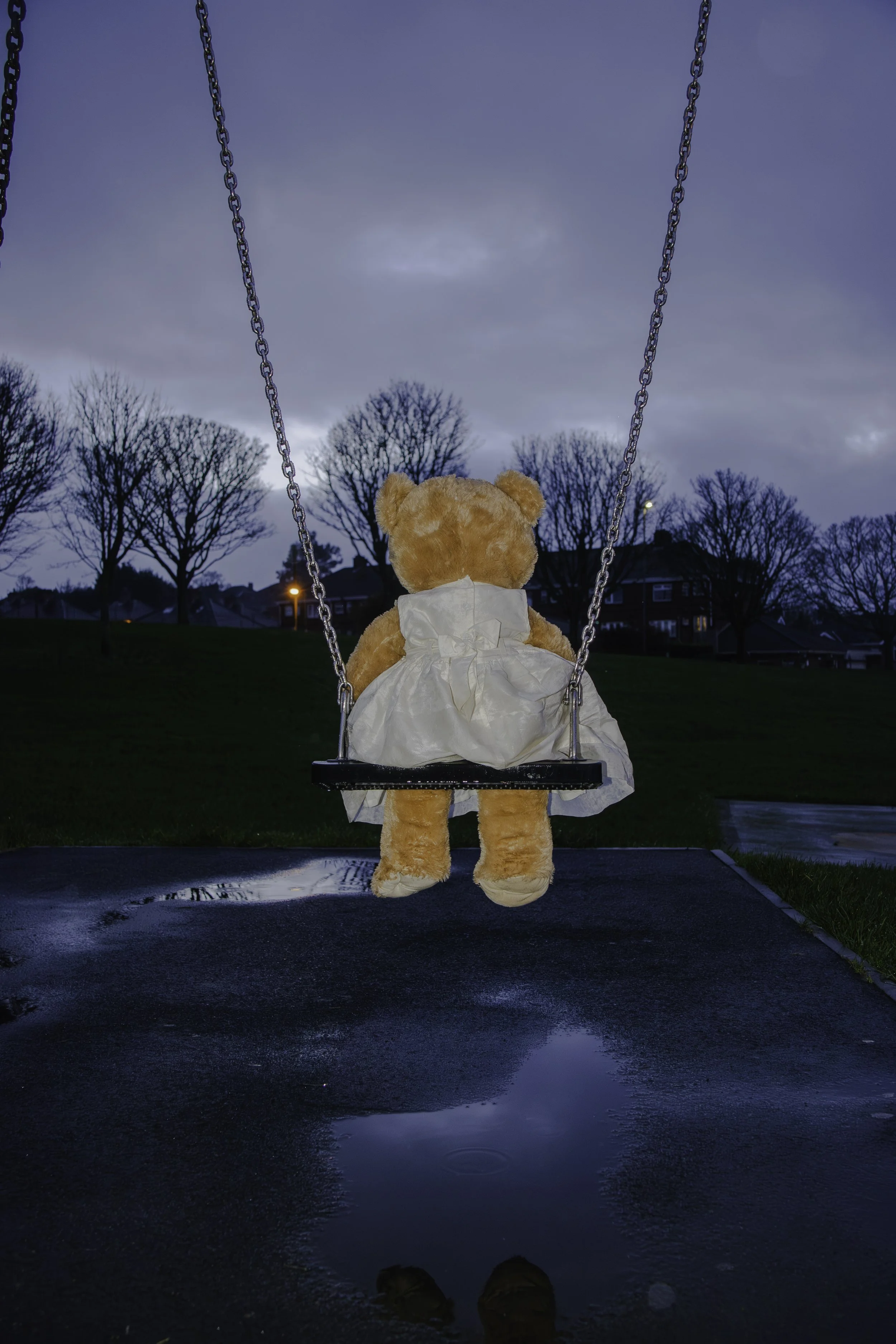 A stuffed bear wearing a white dress sitting on a swing at dusk, with wet ground and puddles reflecting the bear and the swing, trees without leaves, houses, and a cloudy sky in the background.