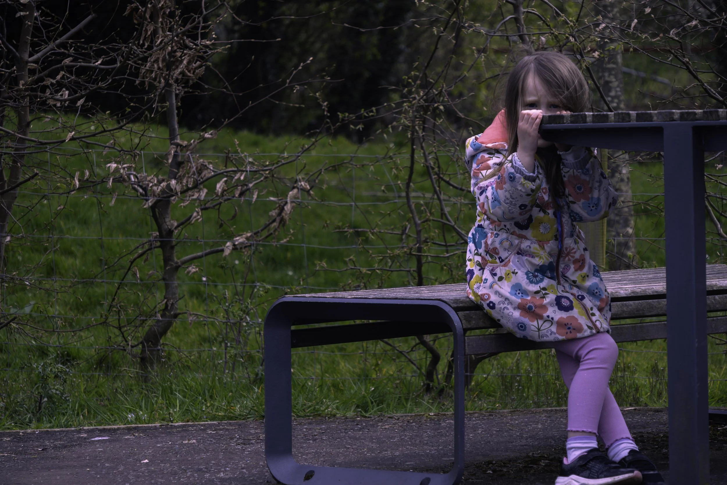A young girl with long brown hair covering part of her face is sitting on a park bench, hiding her face. She is wearing a colourful floral jacket, pink pants, and dark shoes. The background features leafless trees and green grass.