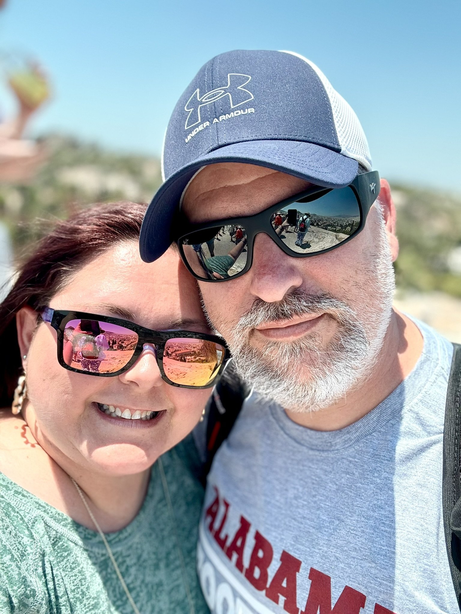 Close-up of a smiling woman with dark hair and sunglasses and a man with gray hair, a beard, and sunglasses, wearing a blue Under Armour cap. An outdoors setting with a clear sky is reflected in their sunglasses.