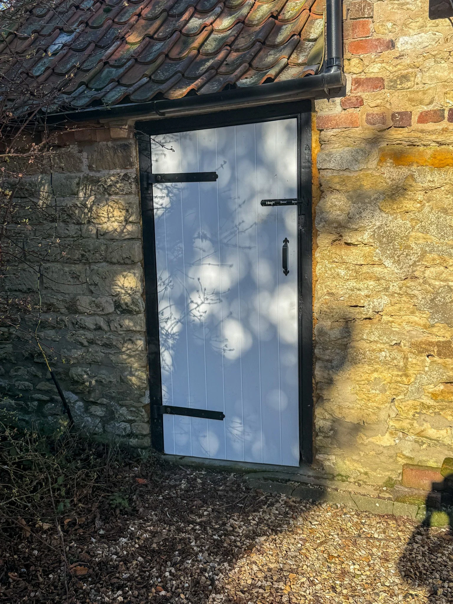 A small door with a black frame and black hardware, attached to a stone wall and a tiled roof. The door's surface reflects the cloudy sky and trees.