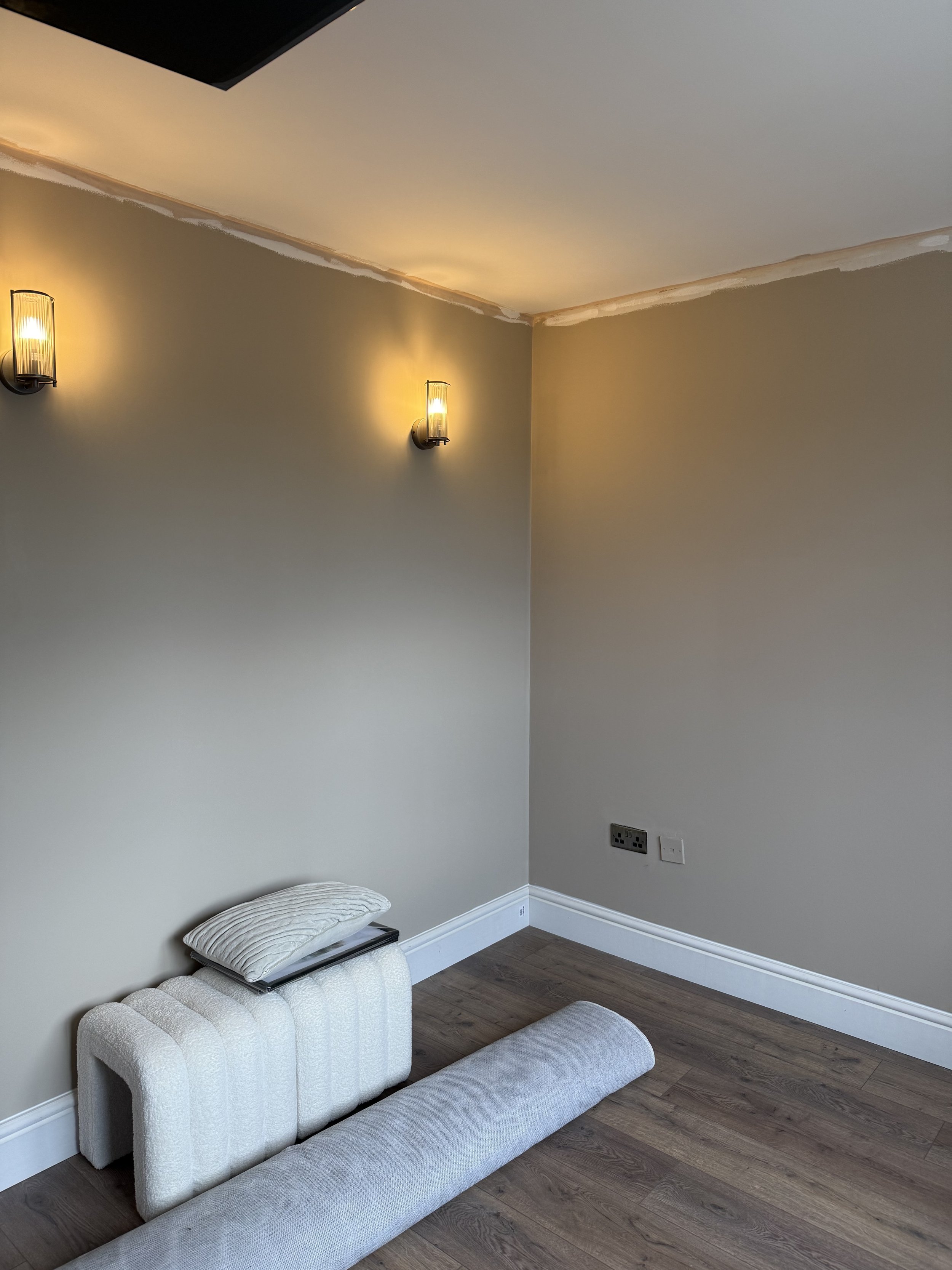 A corner of a room with two wall-mounted lights, a gray couch, and a DIY headboard with cushions on it. The room has light-colored walls, hardwood flooring, and some electrical outlets on the wall.