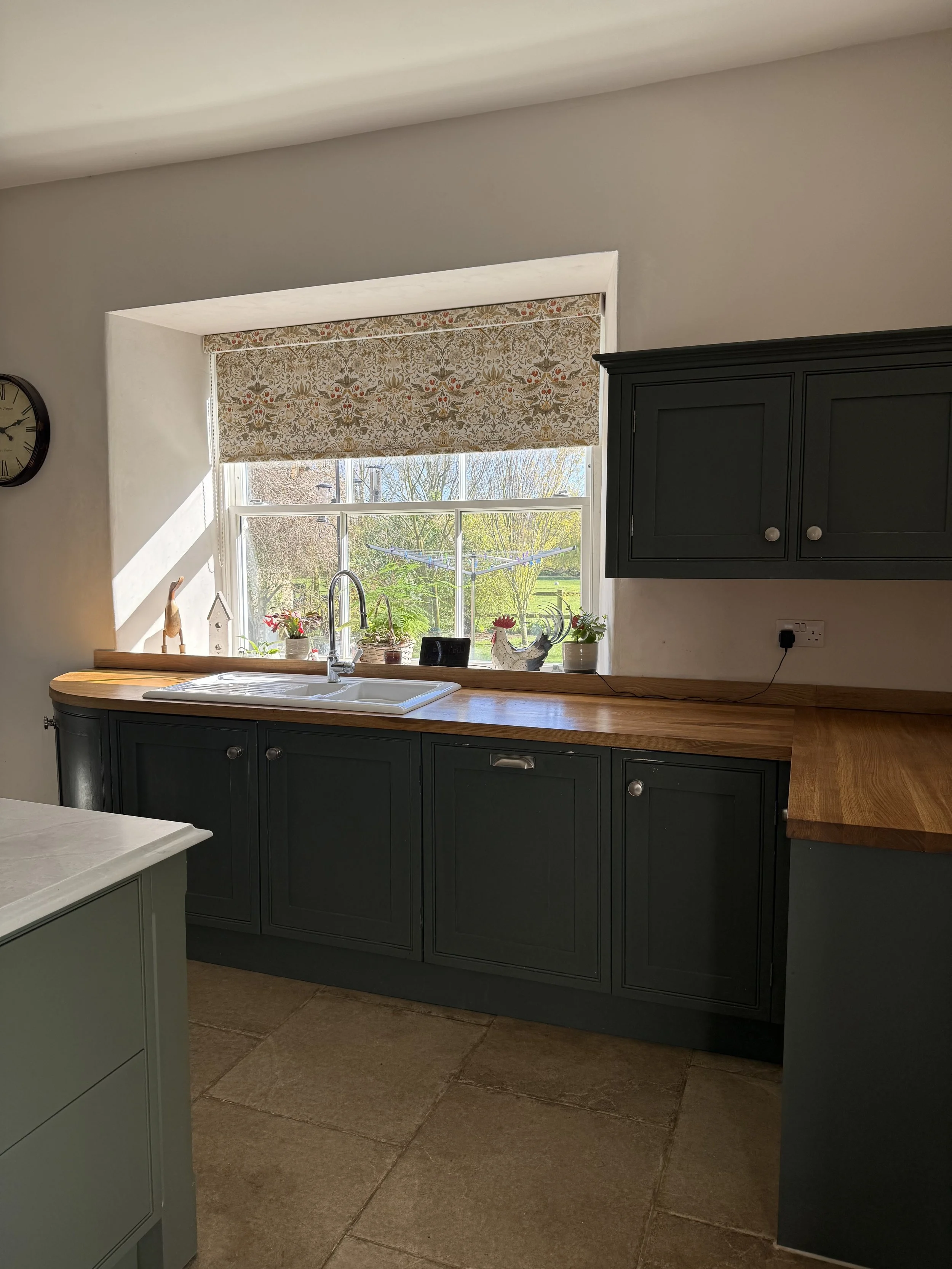 View of a kitchen with a window above the sink, dark green cabinets, a wooden countertop, and decorative plants and figurines on the windowsill.