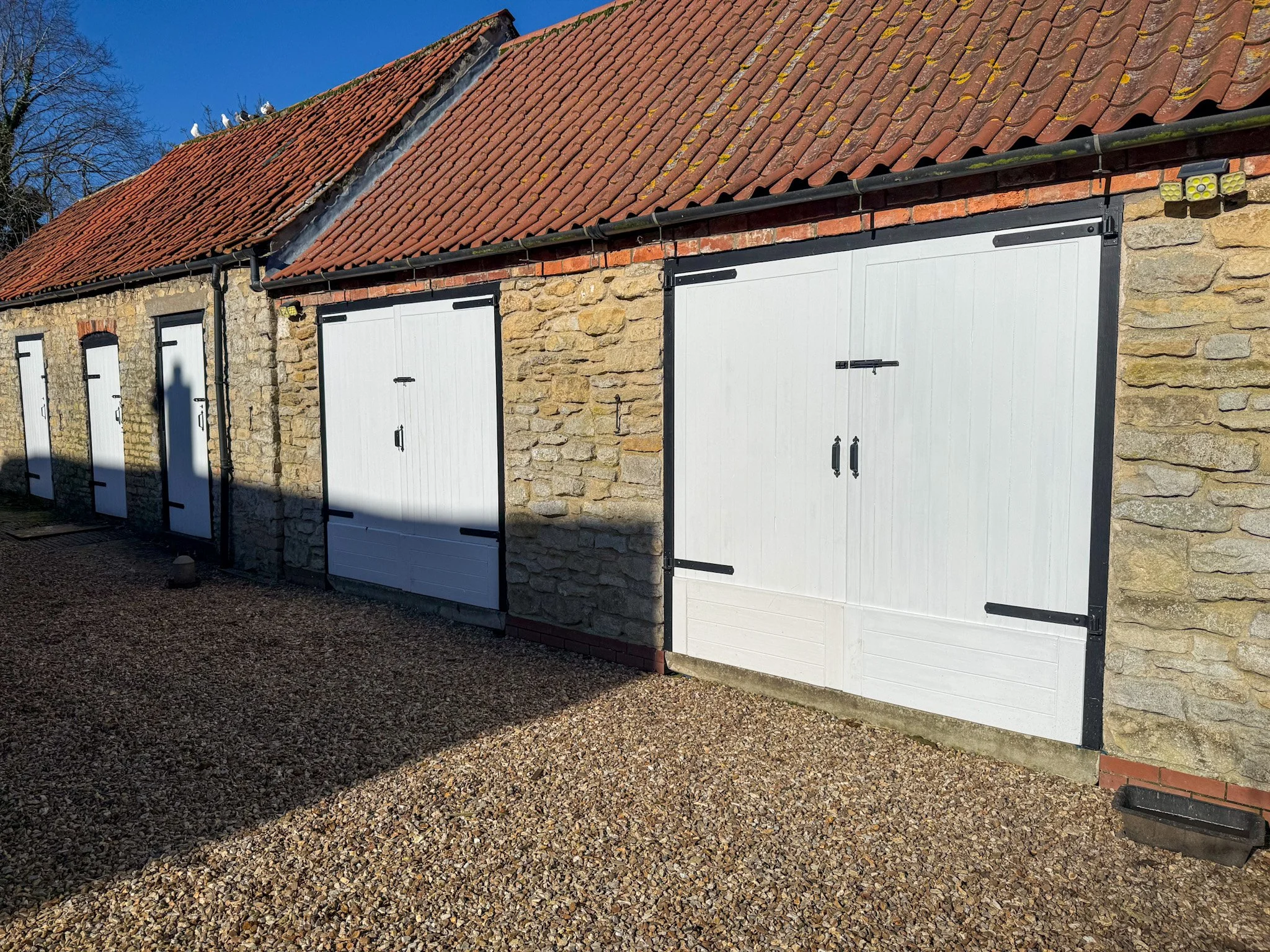 A row of small stone garage buildings with white doors and black trim, under a red-tiled roof, on a gravel driveway with shadows cast on the wall.