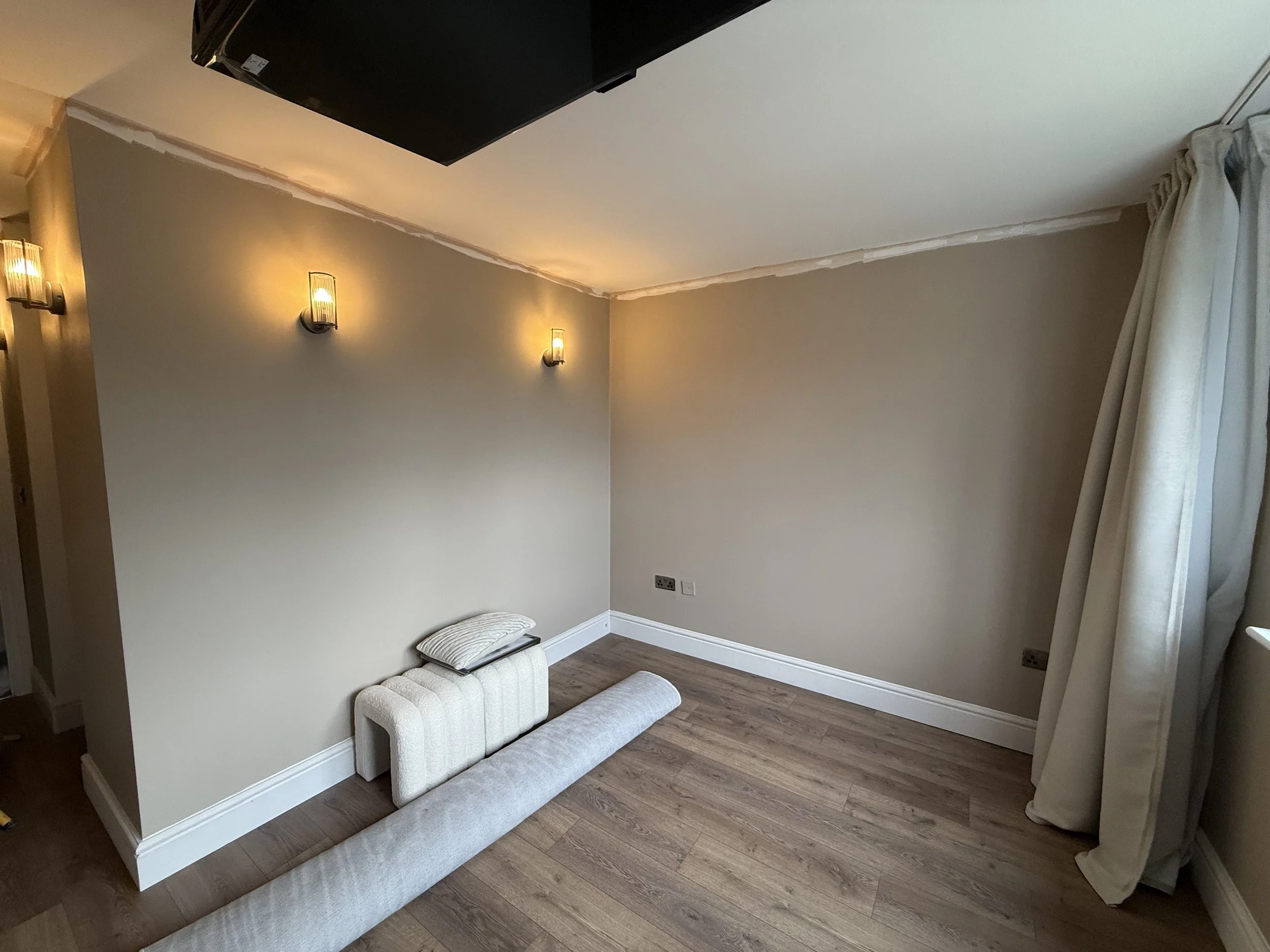 A partial view of a room with a beige wall, two wall-mounted light fixtures, a portion of a window with beige curtains, and a wooden floor. There is a pillow and a long cylindrical cushion on the floor.
