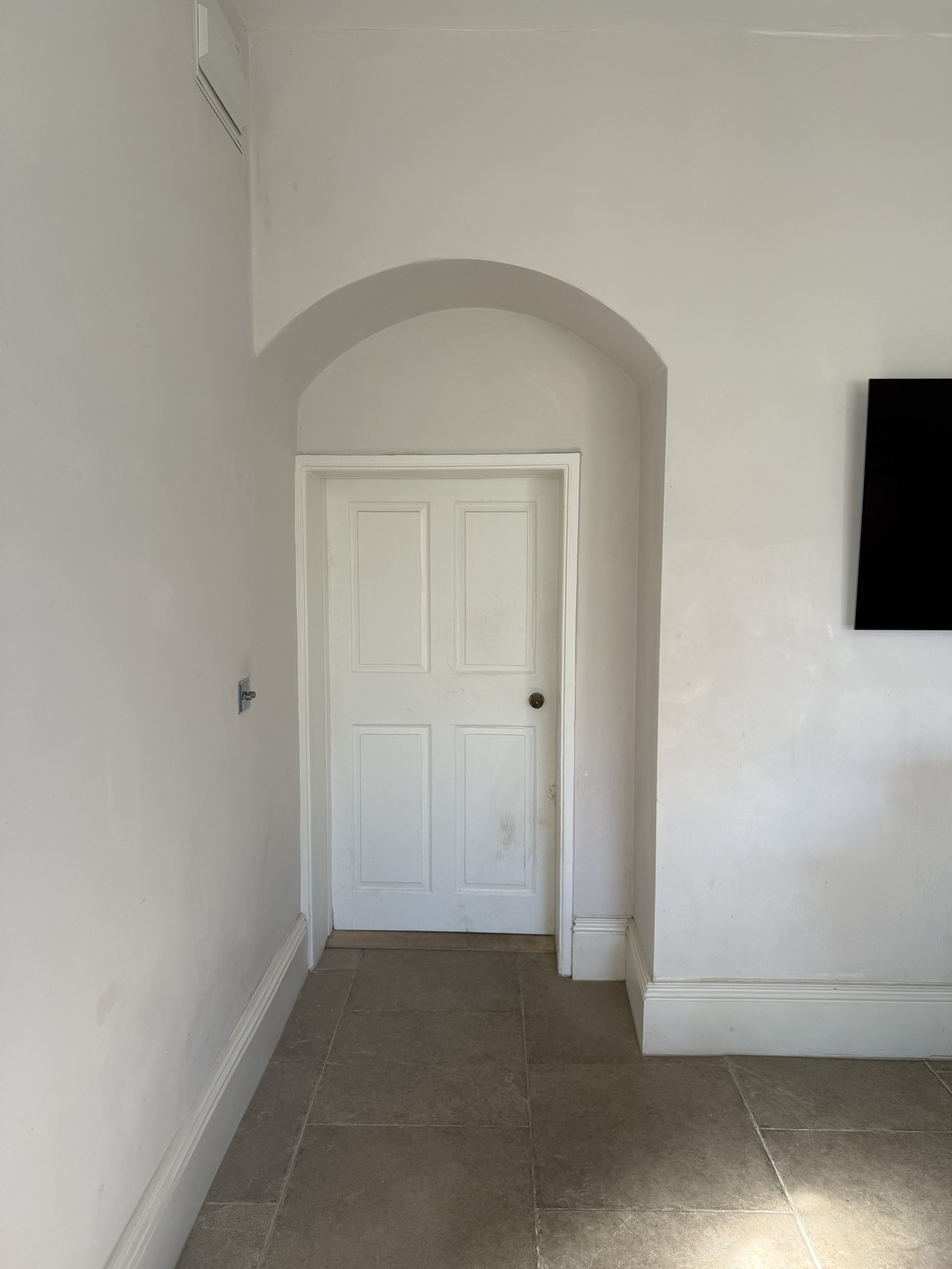 Interior view of a room with a white door, arched doorway, tiled floor, and a wall-mounted TV.
