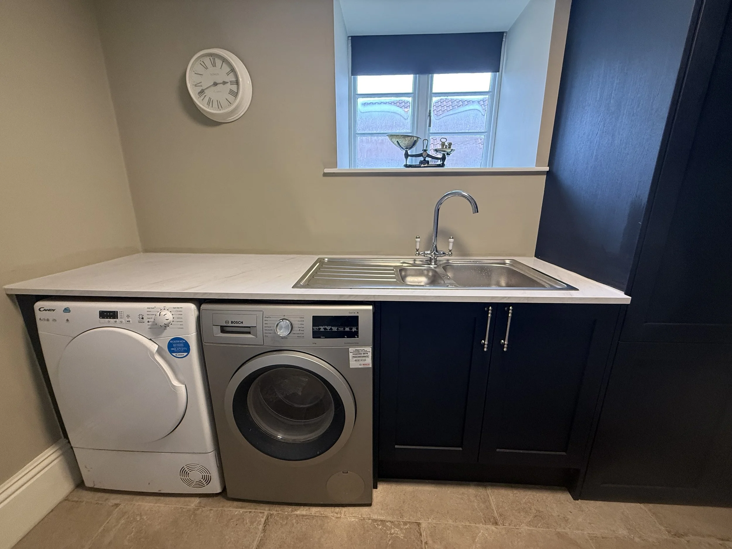 Laundry room with a white washing machine, gray dryer, sink, black cabinets, window with blue roller blind, and a clock on beige wall.