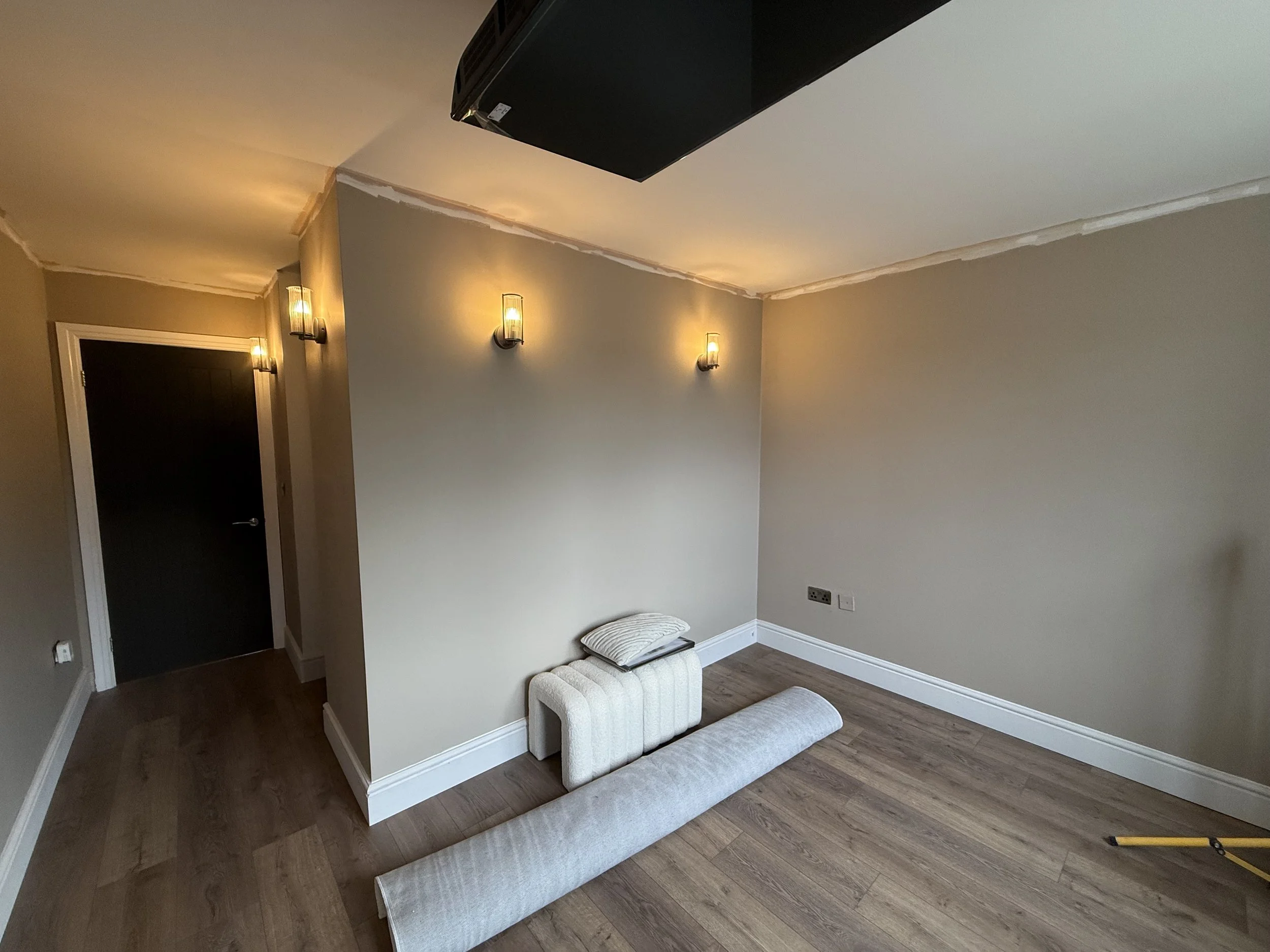 Interior of a room with beige and gray walls, three modern wall sconces, a black ceiling-mounted projector, a white small ottoman, a beige upholstered bench, and hardwood flooring.