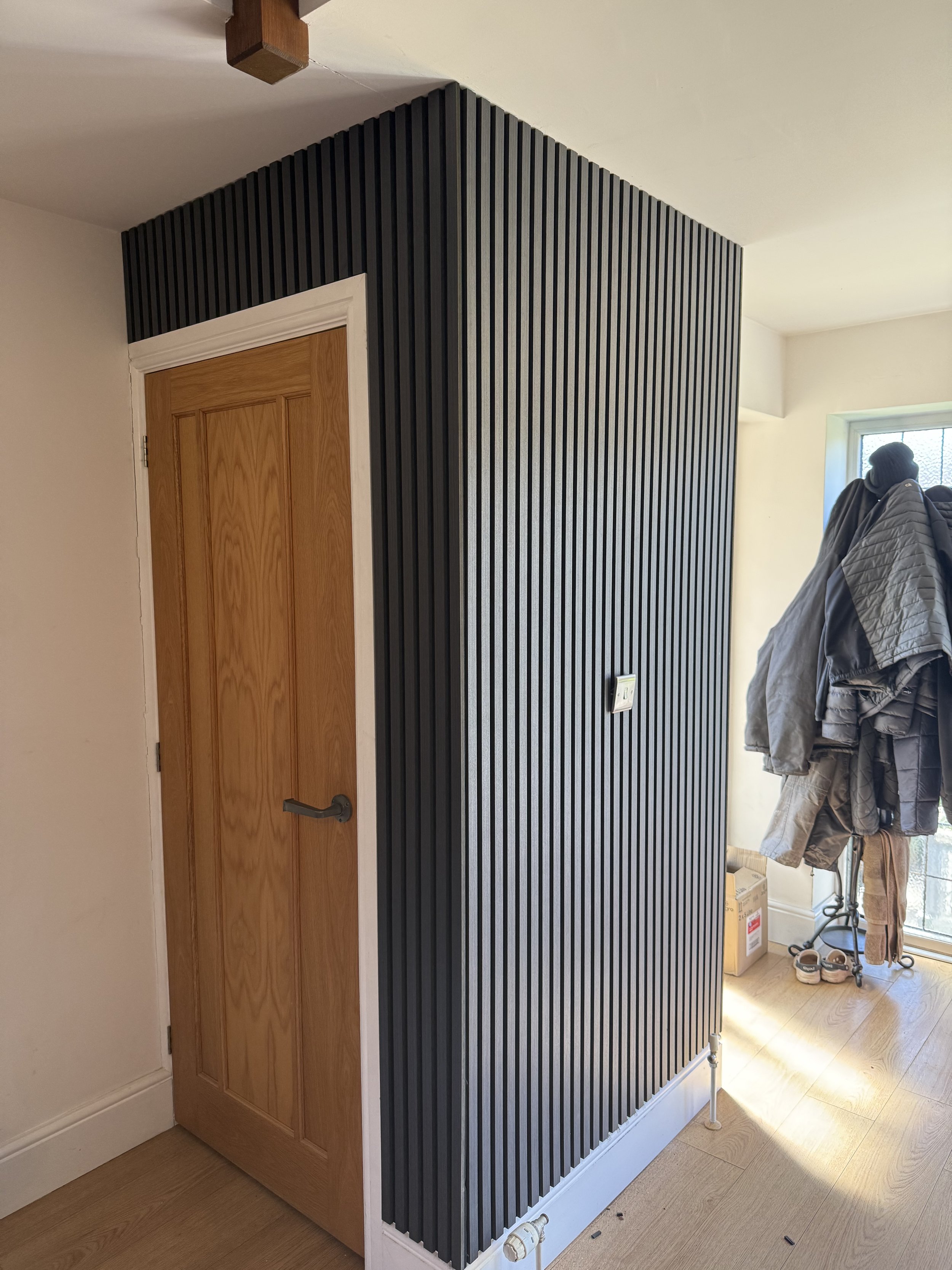 A room with a wooden door, a black slatted wall, coat hooks with jackets and shoes, a window, and some boxes.