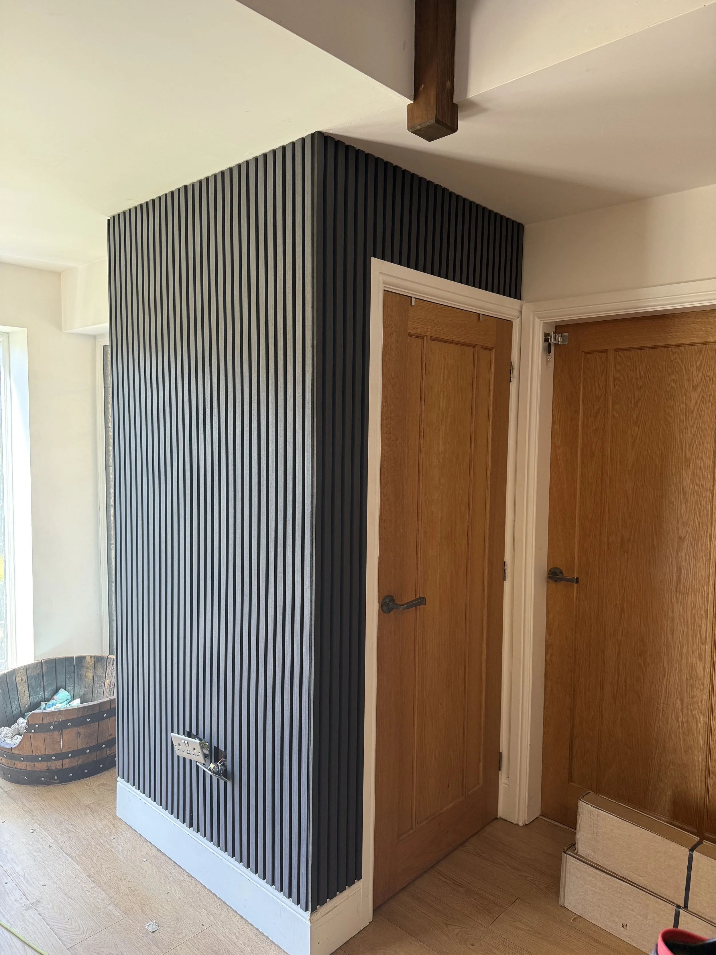 Interior corner of a room with a wooden door, a black vertical slatted panel, a small floor vent, and a hot tub in the background.