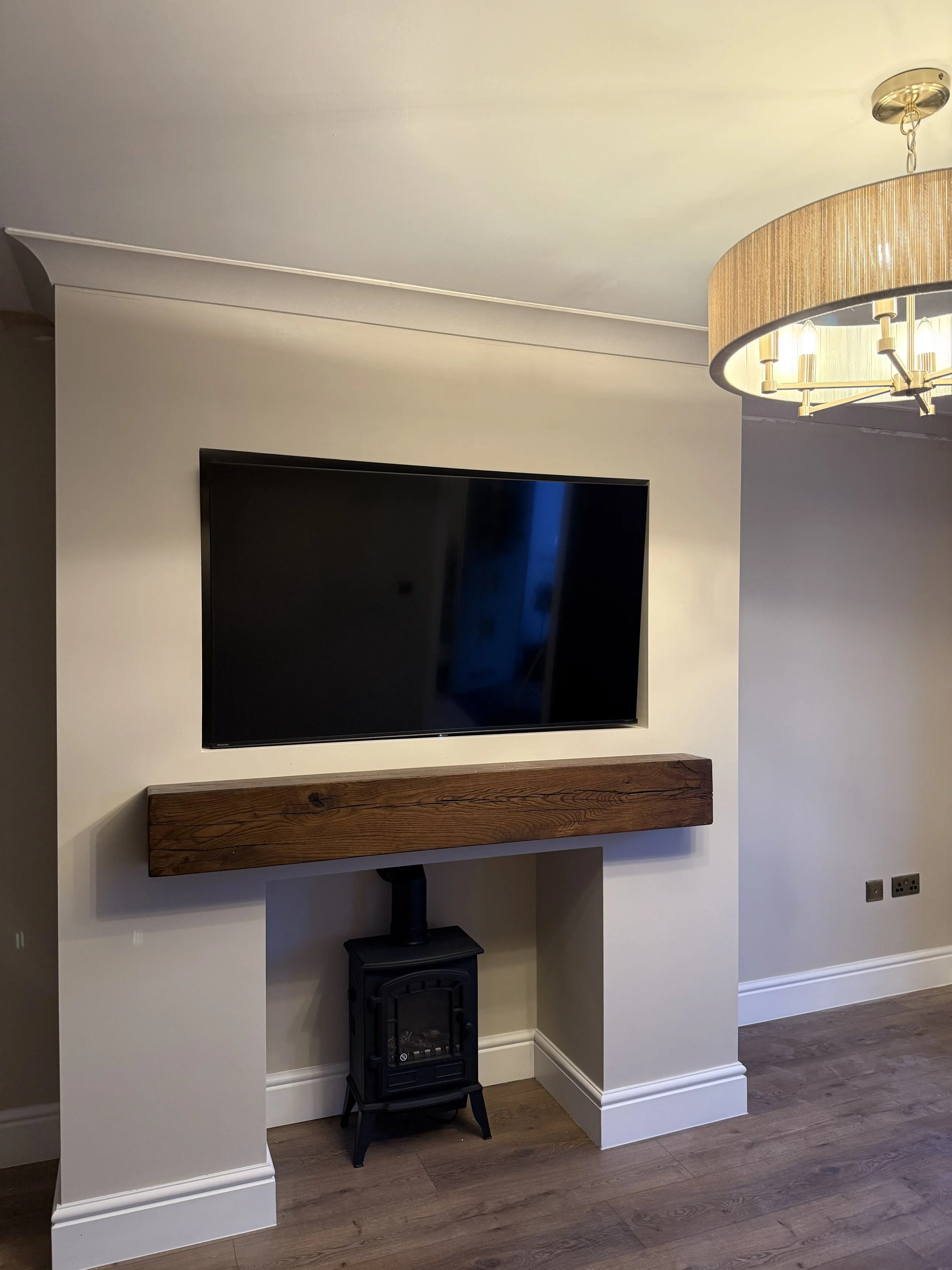 Living room with a mounted flat-screen TV on the wall above a wooden shelf. A small black stove with a chimney pipe is on the floor beneath the shelf. A ceiling light fixture with a beige lampshade is visible in the upper right corner. The room has l