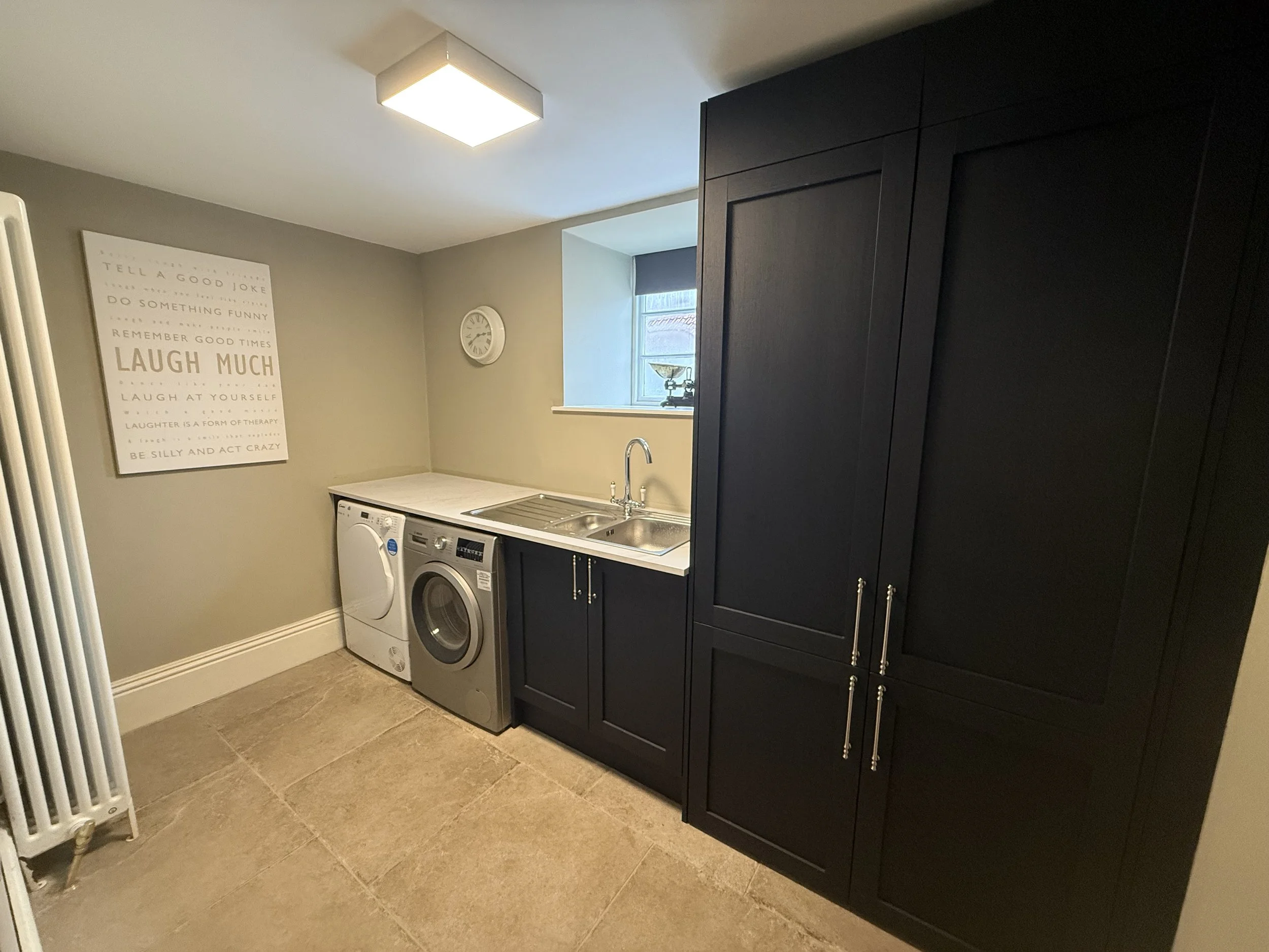 Laundry room with beige tiled floor, gray walls, black cabinetry, washing machine and dryer, stainless steel sink, window, clock, and wall art.