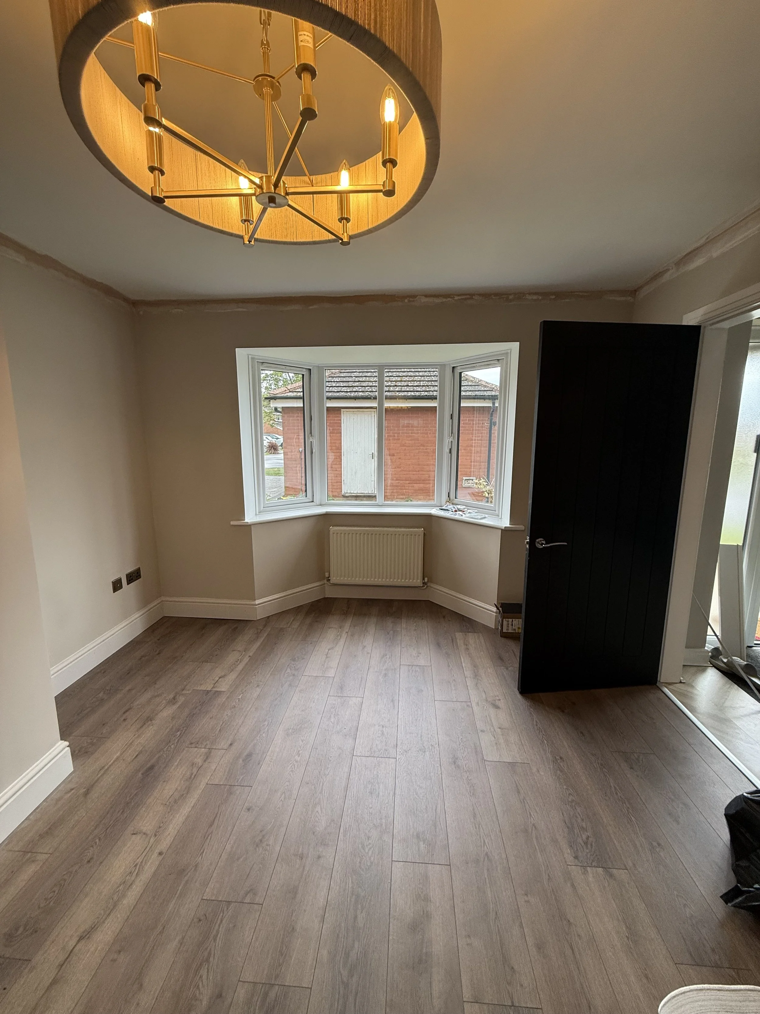 Empty living room with wooden flooring, beige walls, a bay window with a white frame, and a modern gold chandelier lighting fixture on the ceiling.