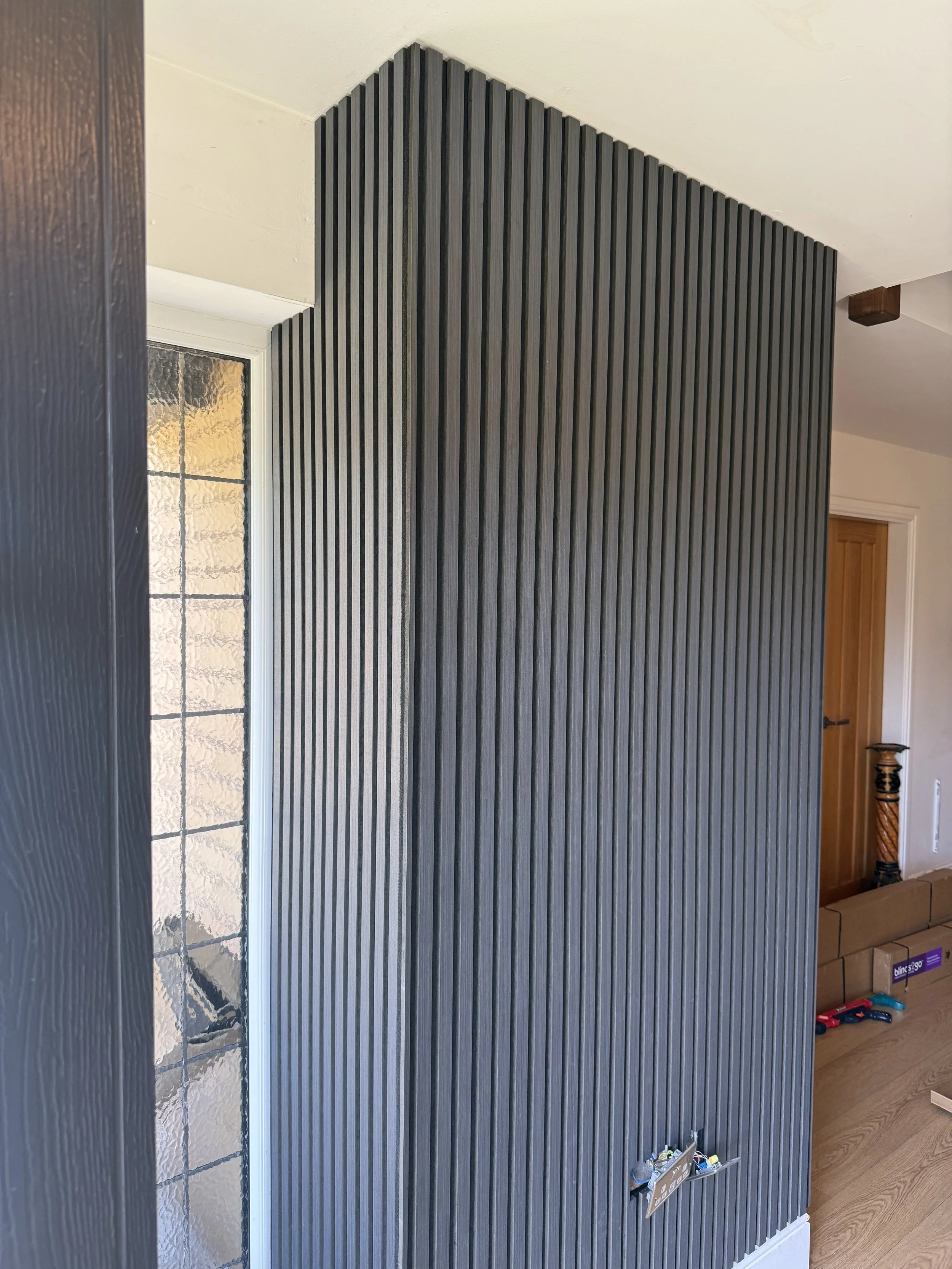 Interior wall with vertical black slats, electrical outlets, and a nearby window with textured glass, in a modern home.