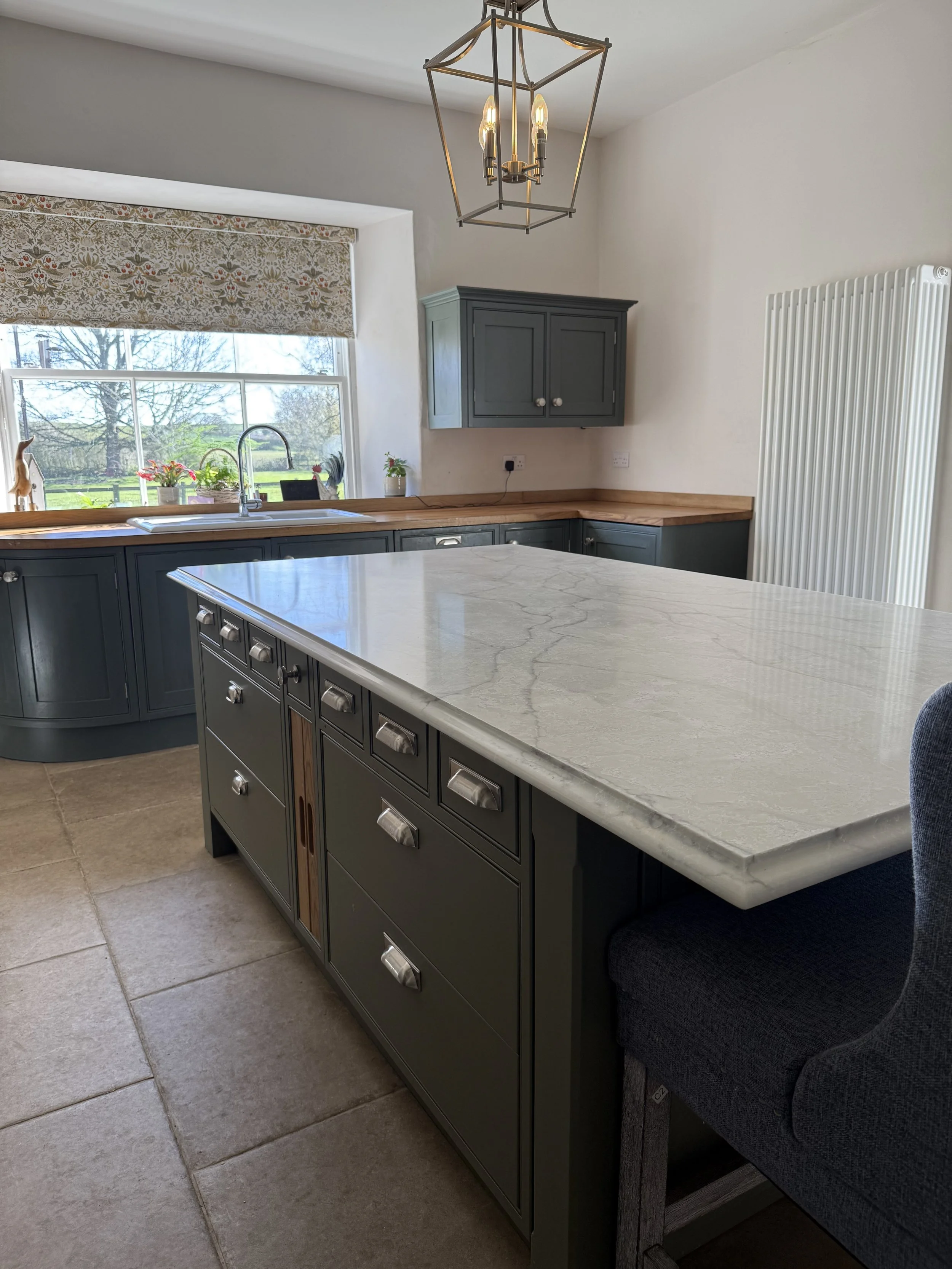 Modern kitchen with gray cabinets, a large island with a marble countertop, a window with a floral valance, a black table lamp, and a brass geometric pendant light hanging from the ceiling.