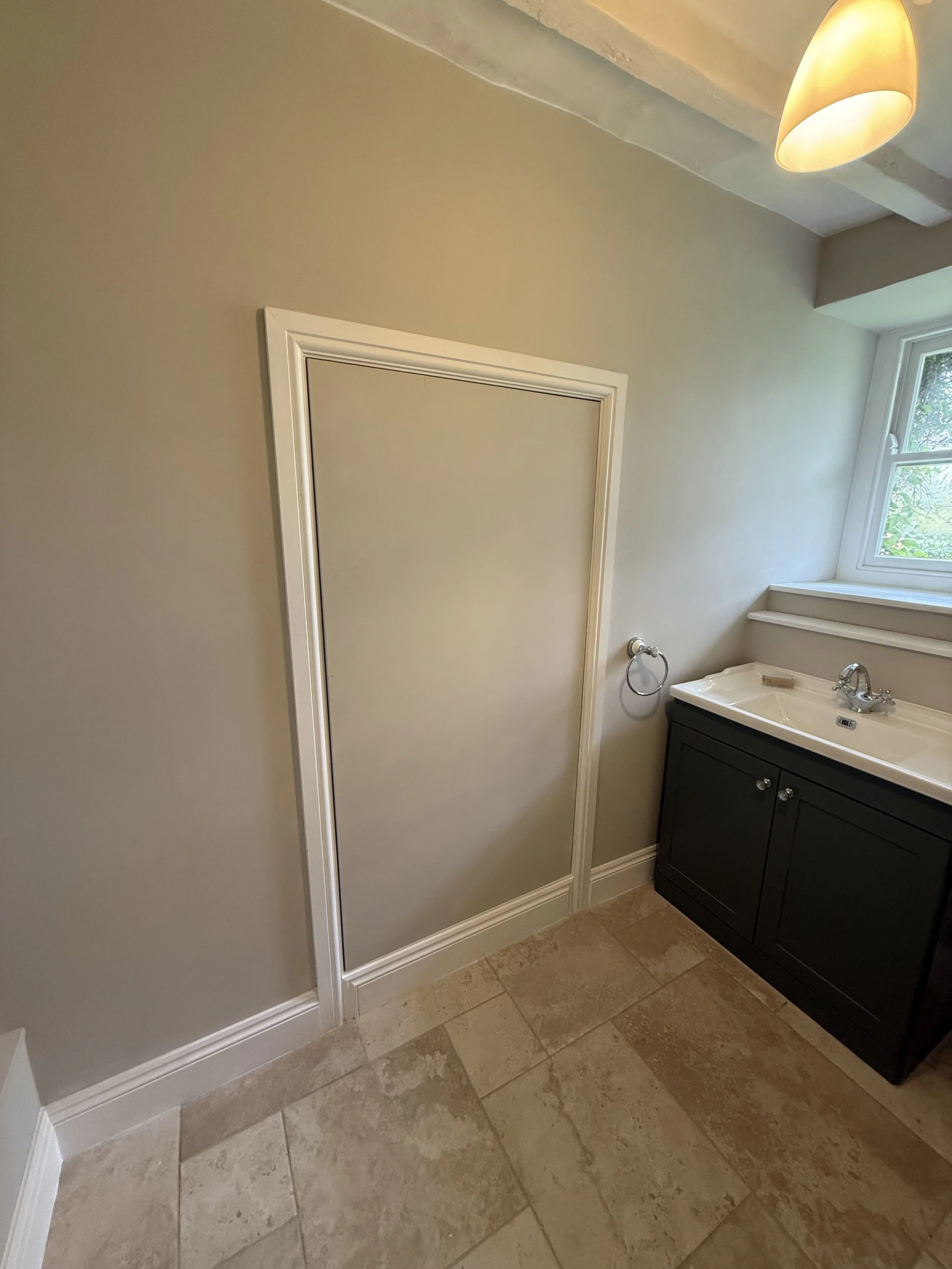 A small bathroom with beige tiled flooring, a wall-mounted mirror, a dark-colored vanity with a white sink, a window with natural light, and a ceiling light fixture.