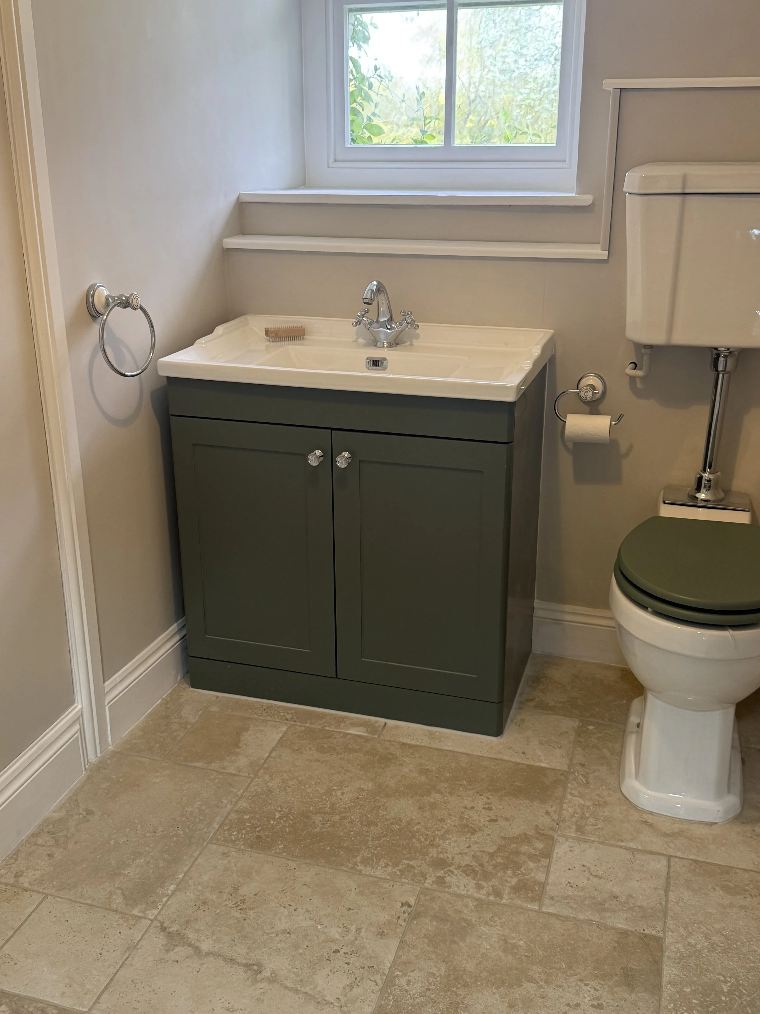 Small bathroom with beige tiled floor, white walls, a green vanity with a sink, a window above the vanity, a toilet with a green seat cover, and a silver towel ring beside the vanity.