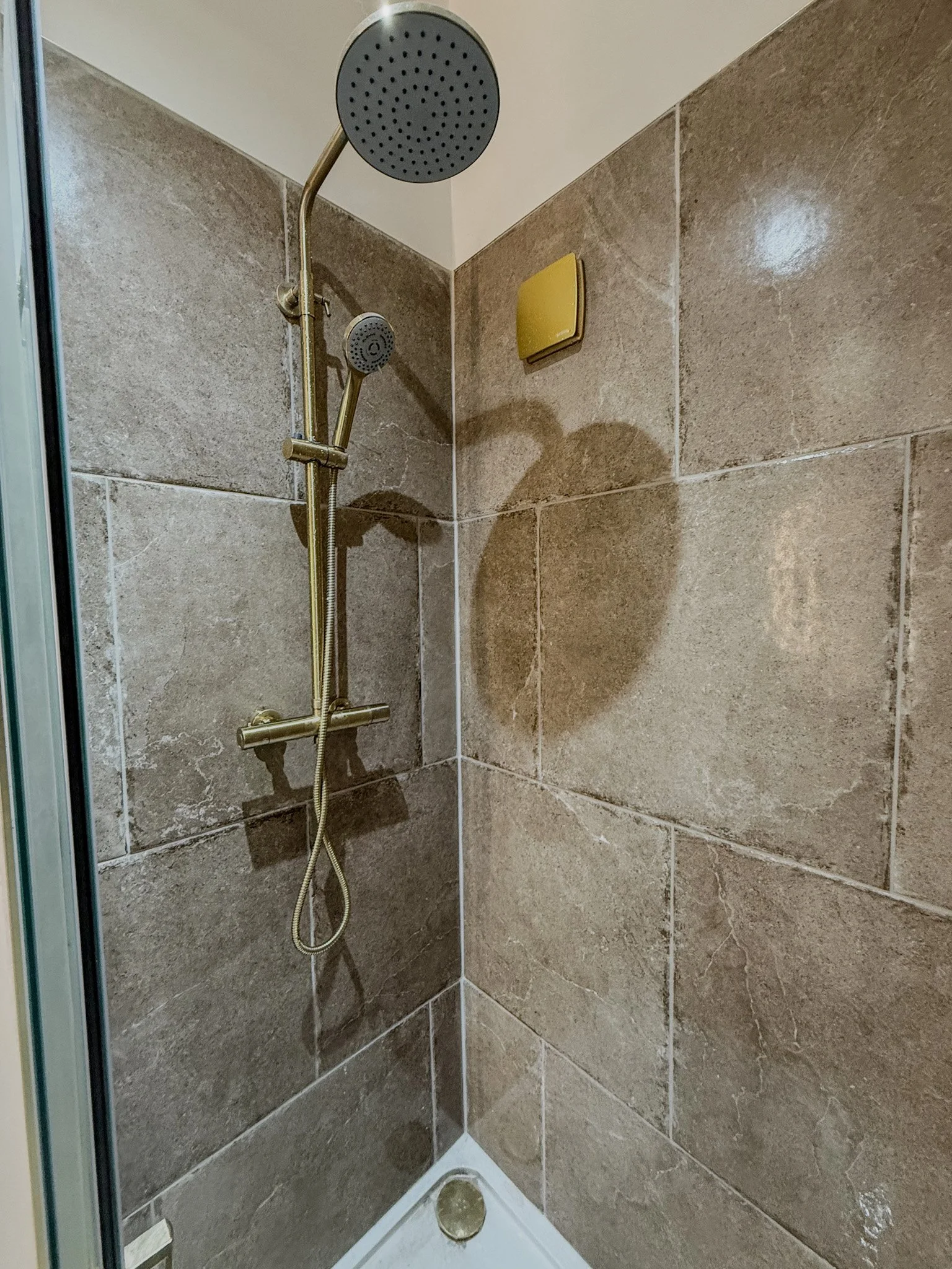 Shower stall with brown tiled walls, a round showerhead, a secondary handheld shower, and a yellow vent on the wall.