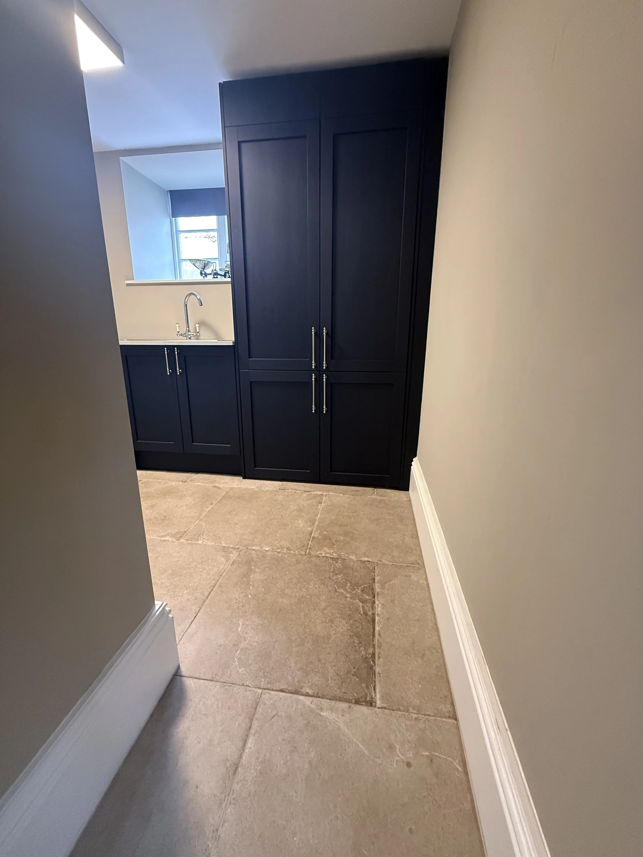 A small section of a kitchen or utility area with beige tile flooring and dark navy blue cabinets, a white countertop, and a window with a roller shade.