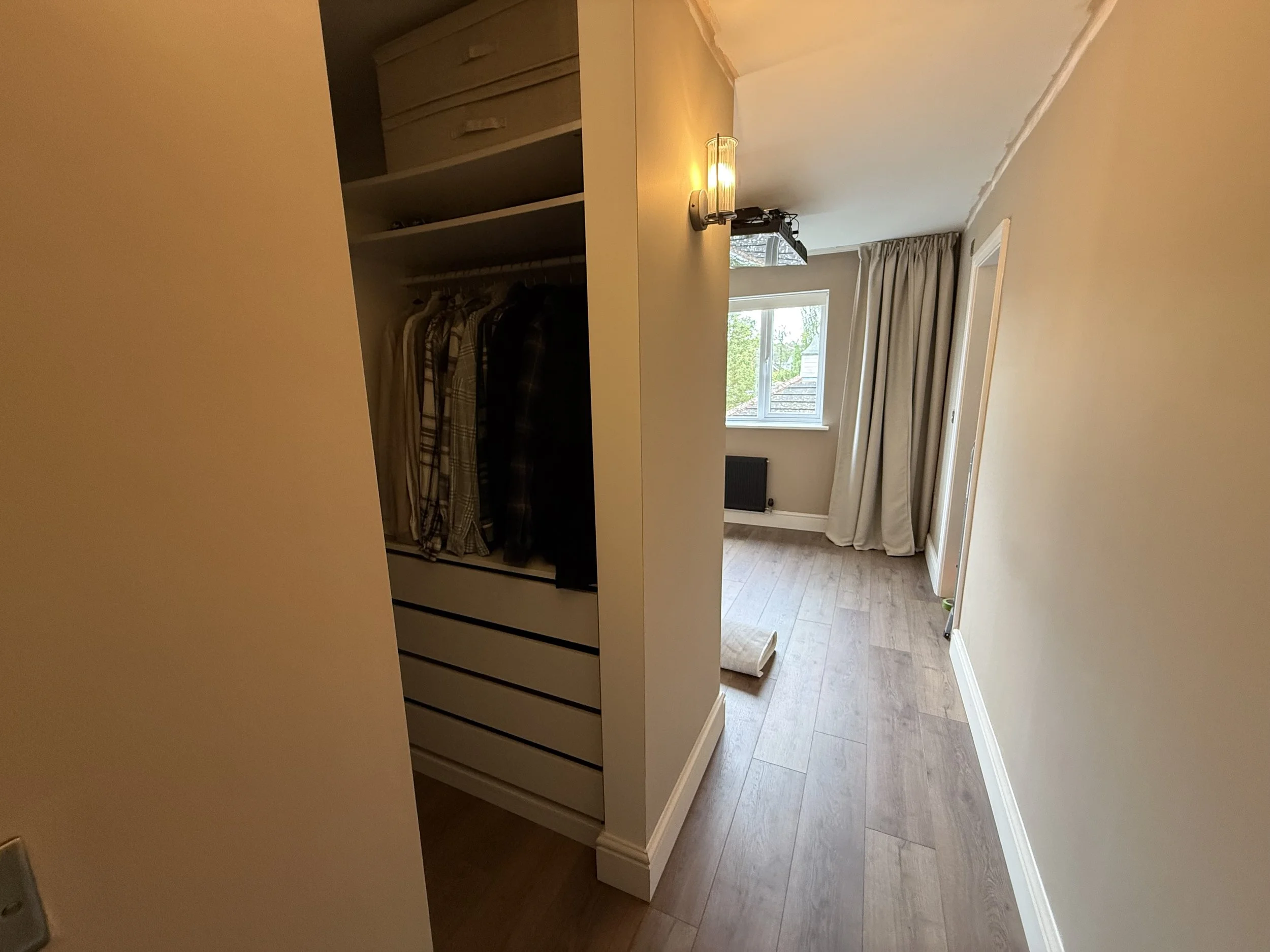 A view of a cozy living room with a window with beige curtains, a radiator, a floor lamp, and a cabinet filled with clothes and shoes.