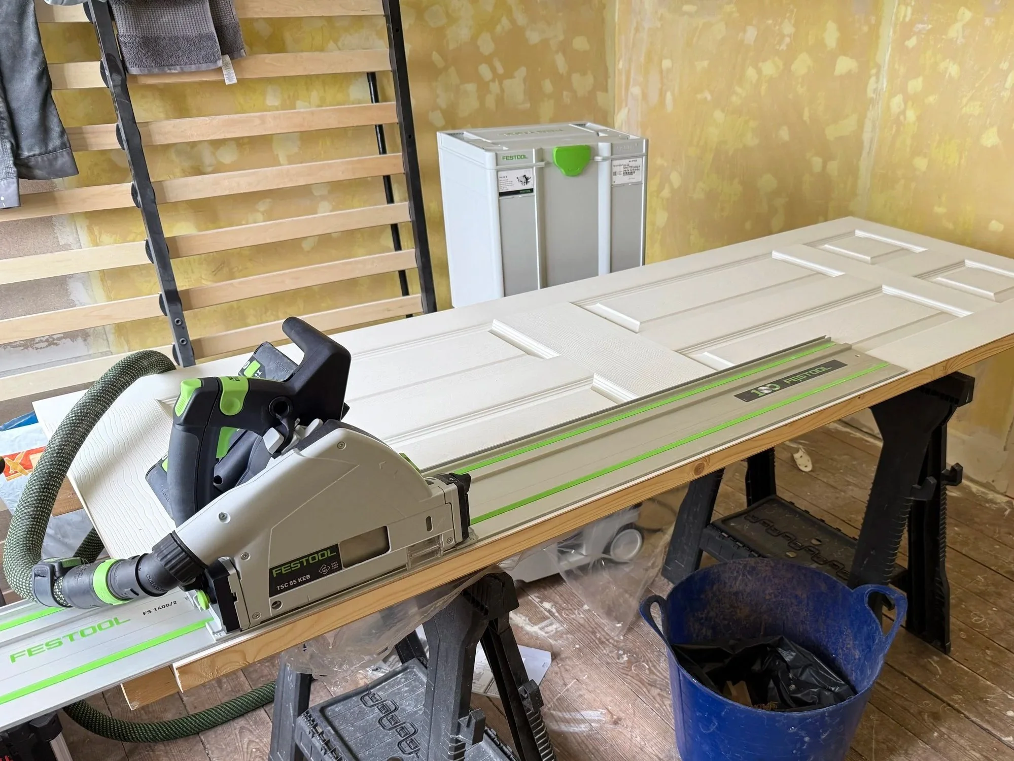 A woodworking workbench with a festool track saw, a large white door panel, a blue trash bin, and a wooden wall with a coat rack.