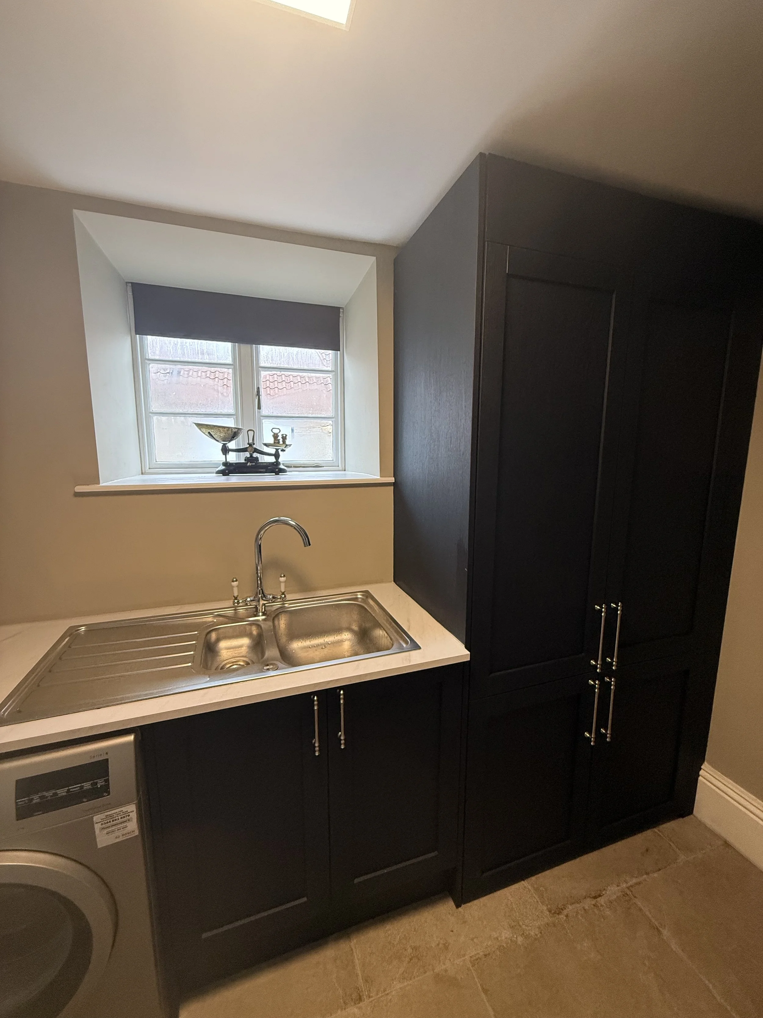 Small laundry area with black cabinets, a stainless steel sink, a window with a gray roller shade, and a washing machine.