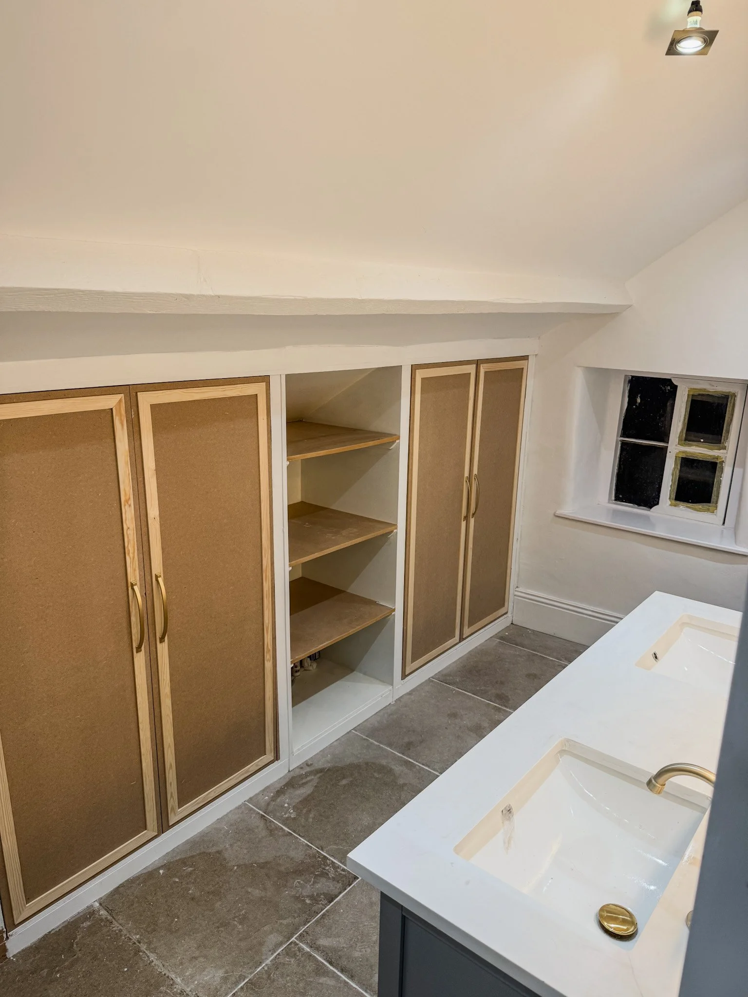A bathroom with two sinks, a small window, and a large cabinet with four doors, some of which are open showing empty shelves, on a gray tile floor.