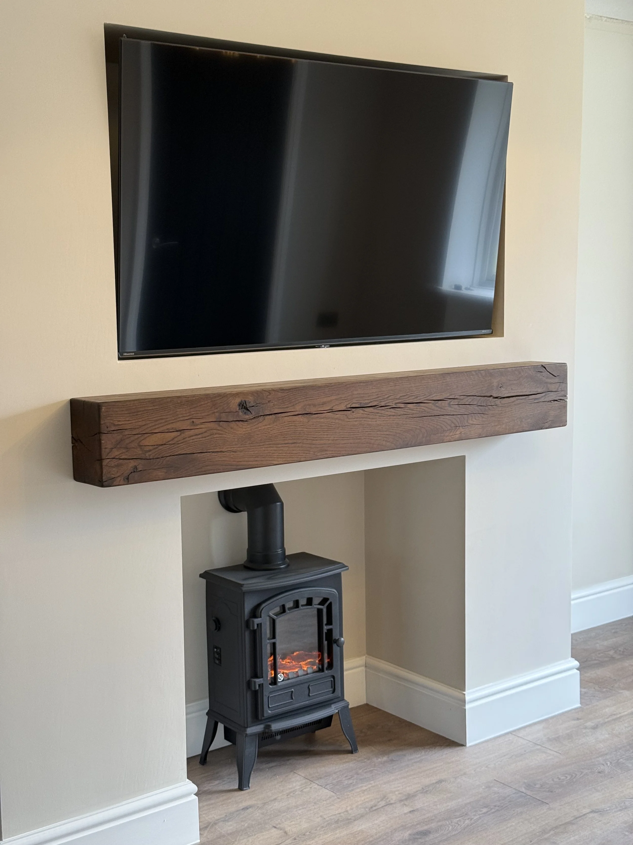 Living room corner with a mounted flat-screen TV above a wood fireplace with a black stove, on a beige wall with white baseboards, and wood flooring.