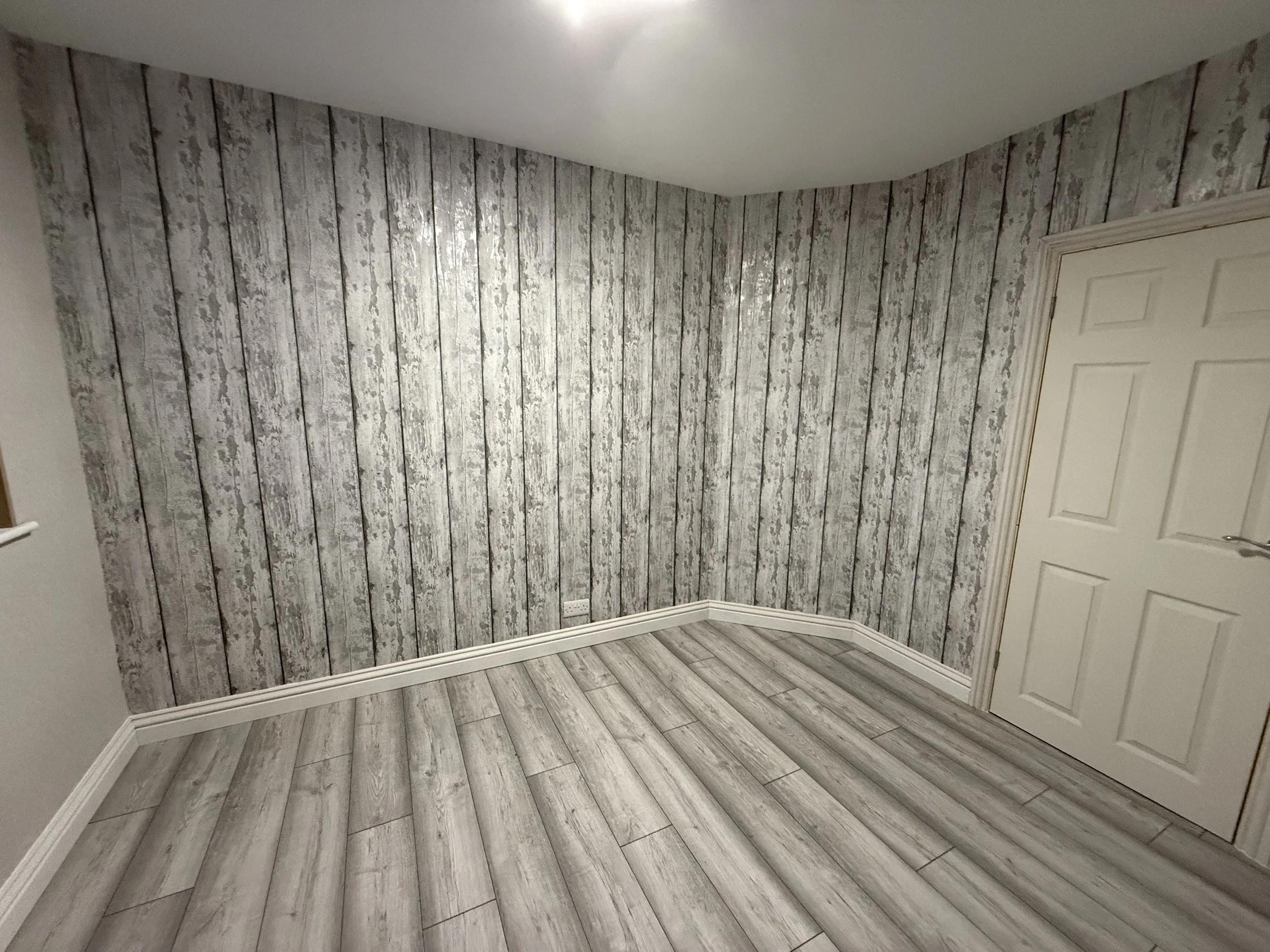 Empty room with wood-patterned wallpaper and matching wood-patterned flooring, white baseboards, a white door with a gold frame, and a ceiling light.